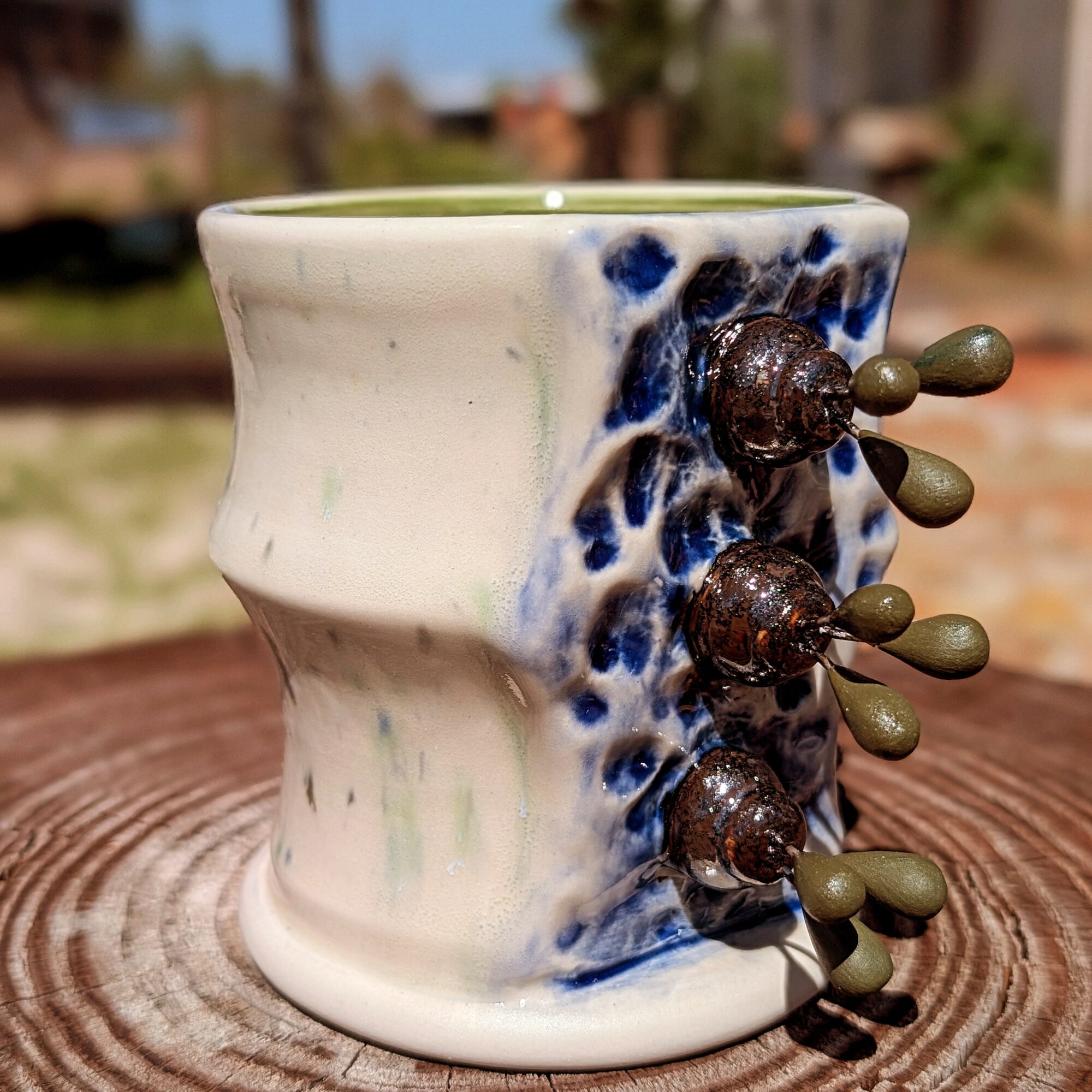 Ceramic mug with blue and white glaze, decorated with small brown and green beads on the side, outdoors on a wooden surface.