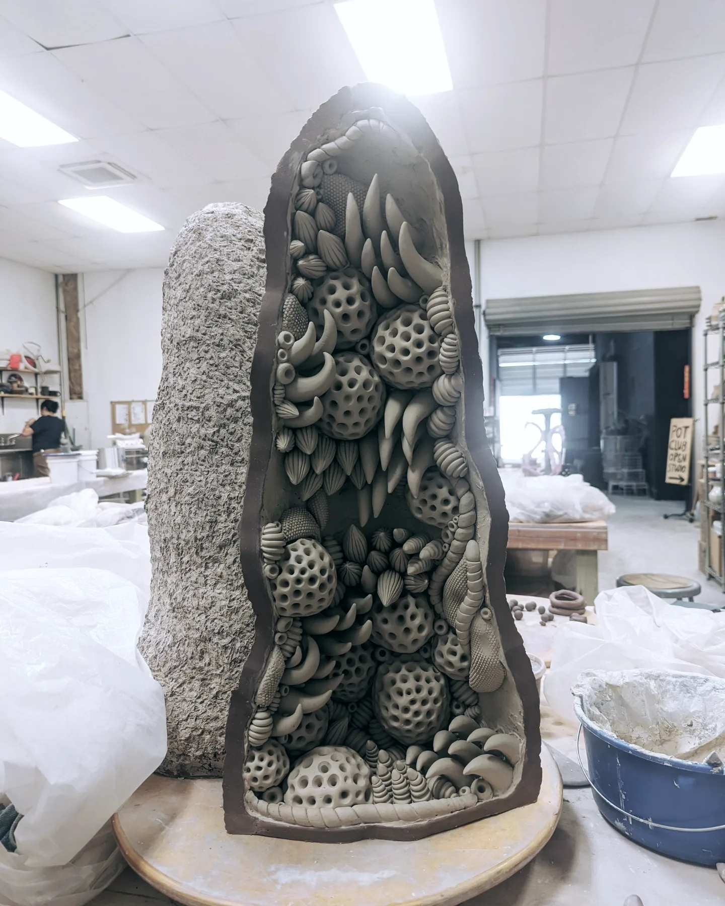 Tall, oval-shaped sculpture filled with various textured, patterned objects, in an indoor workshop setting.