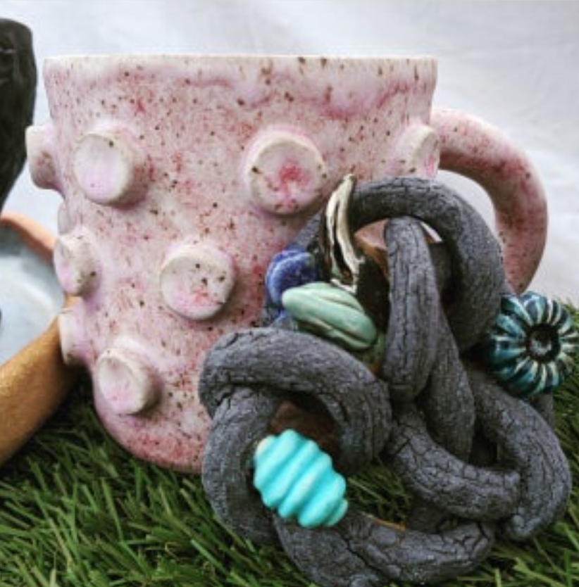 Pink ceramic mug with raised bumps, placed on grass with decorative black and blue twisted objects attached, against a white background.