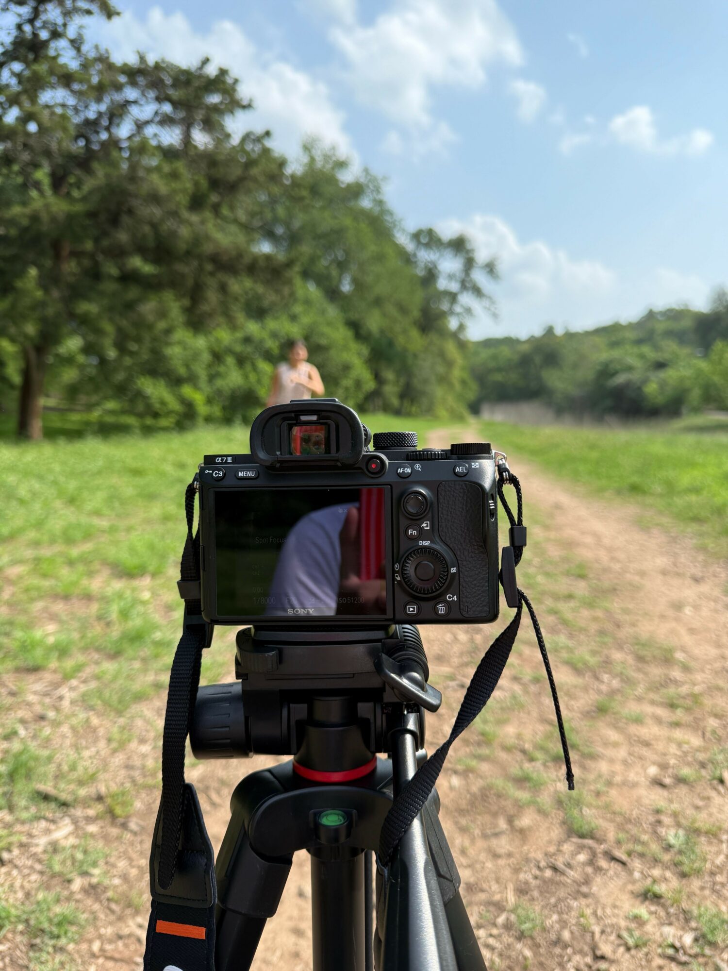 Camera on tripod capturing a person standing outdoors with trees and blue sky in background.