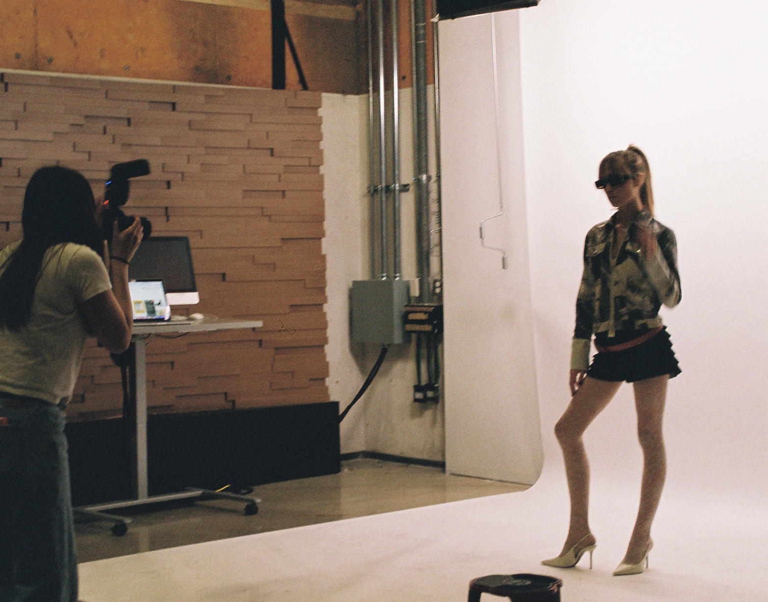 Person in front of a white backdrop, posing with one hand on hip and wearing high heels, while another person takes photos.