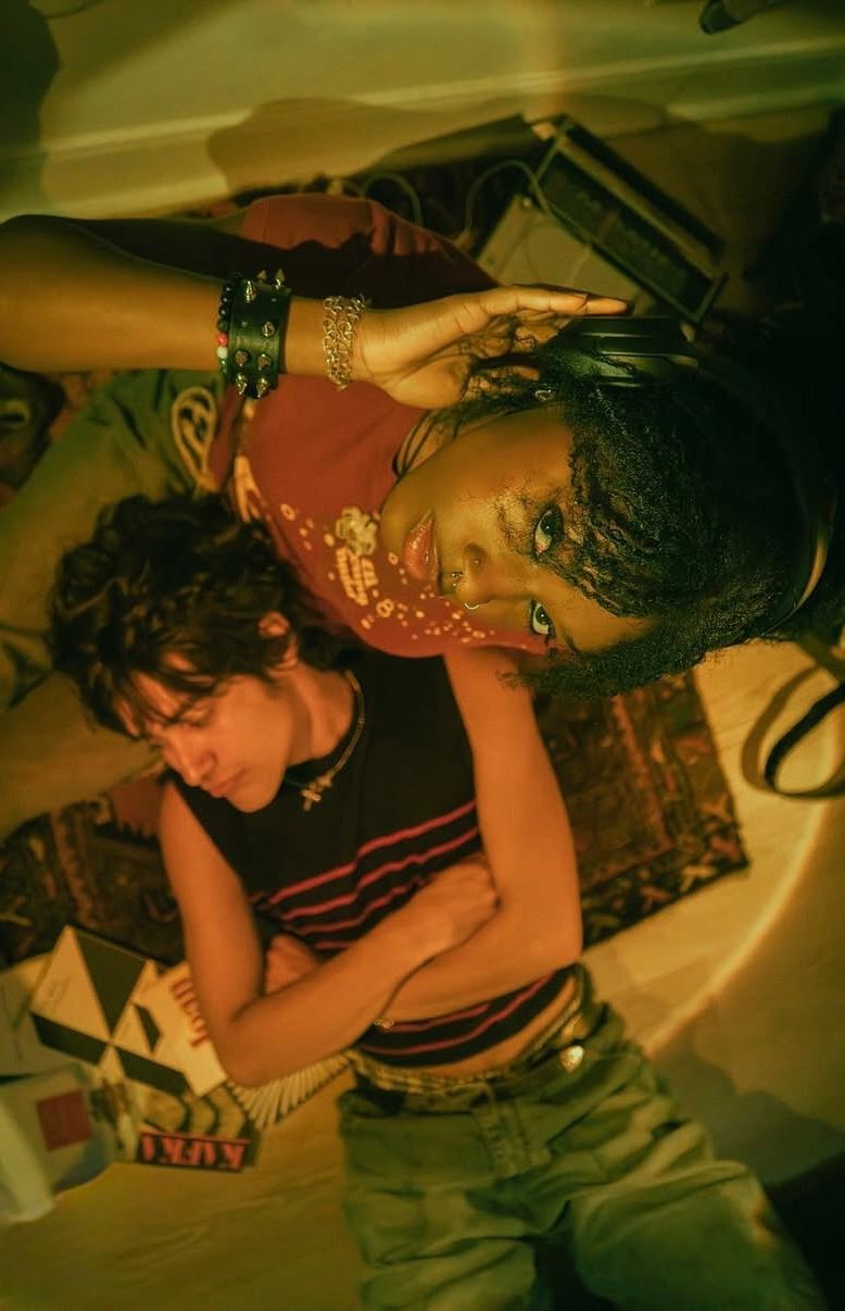 Two women lying on the floor, one with headphones, looking up at the camera, surrounded by books and patterned rugs.