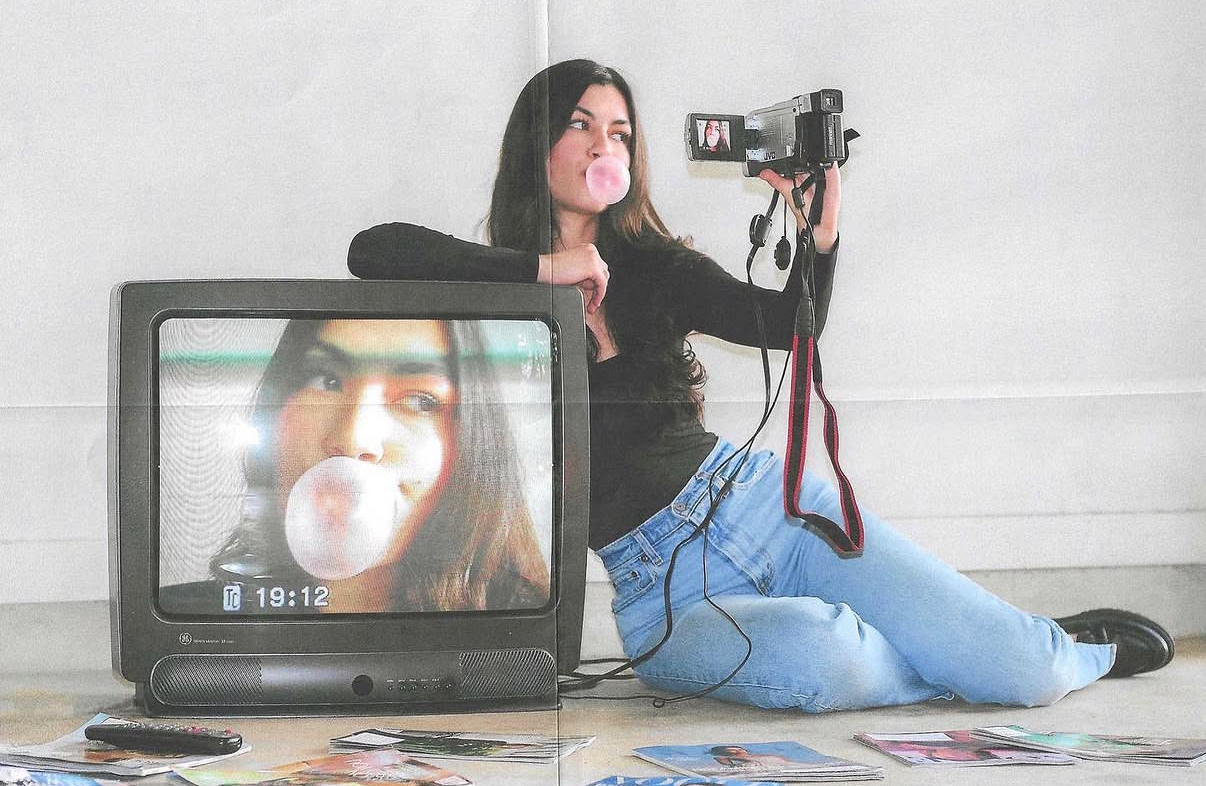 Woman sitting on the floor with a camera, a TV showing her face, and magazines scattered around.