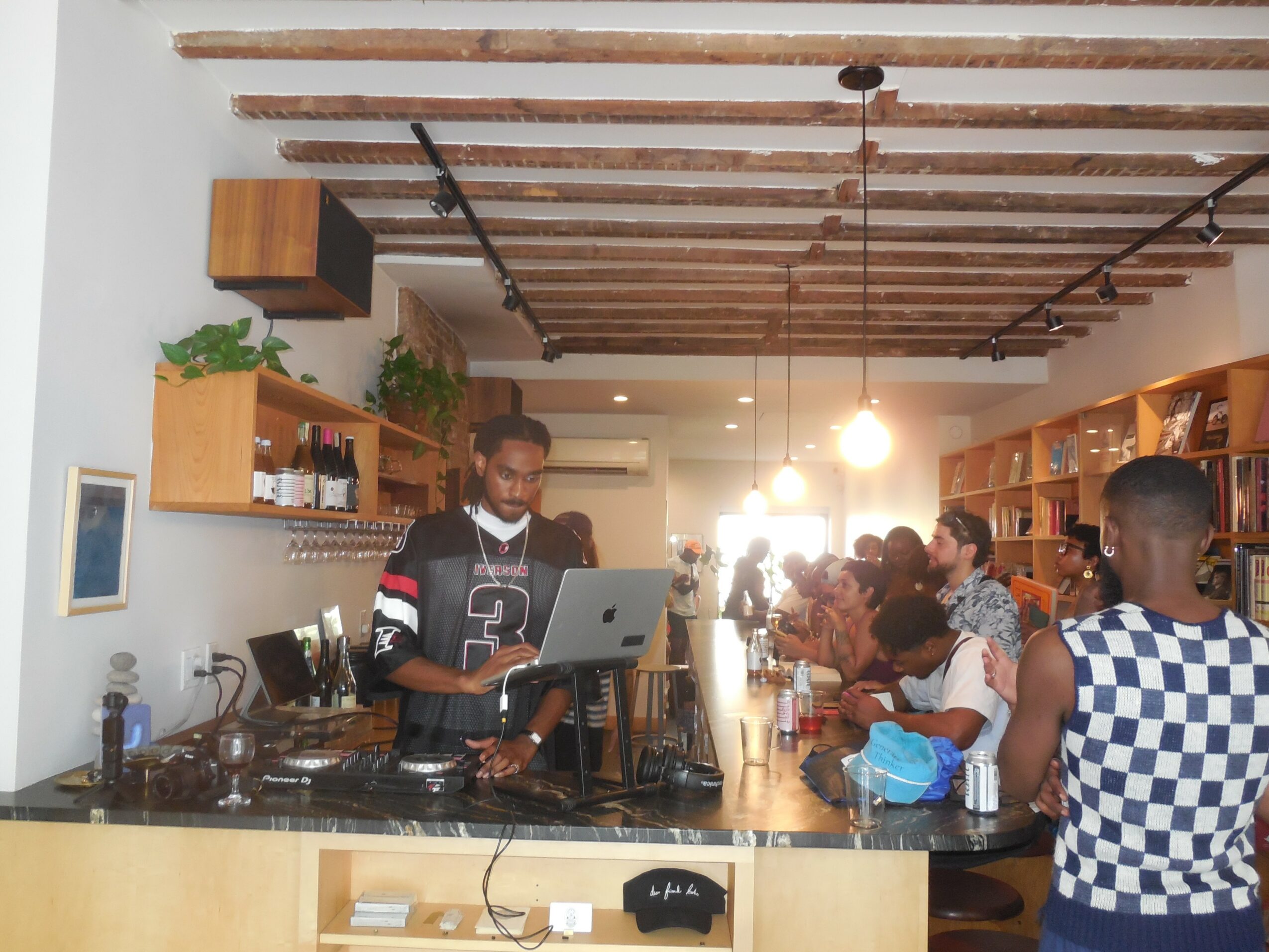 Person working with DJ equipment in a cafe, with customers seated at a bar, wooden ceiling beams, and shelves with bottles.