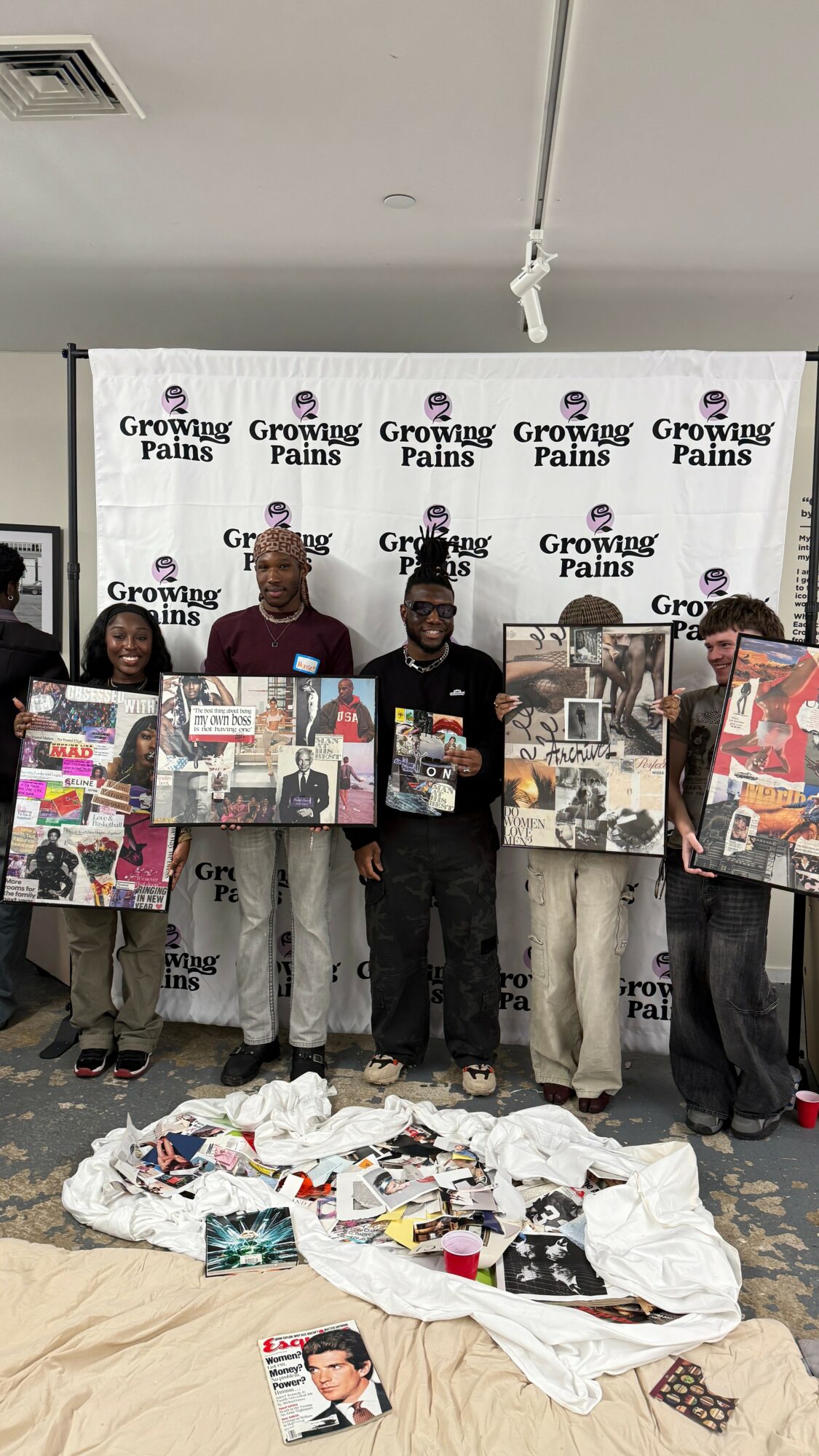 Four people stand indoors holding large posters, with a pile of clothes and items on the floor in front of them.