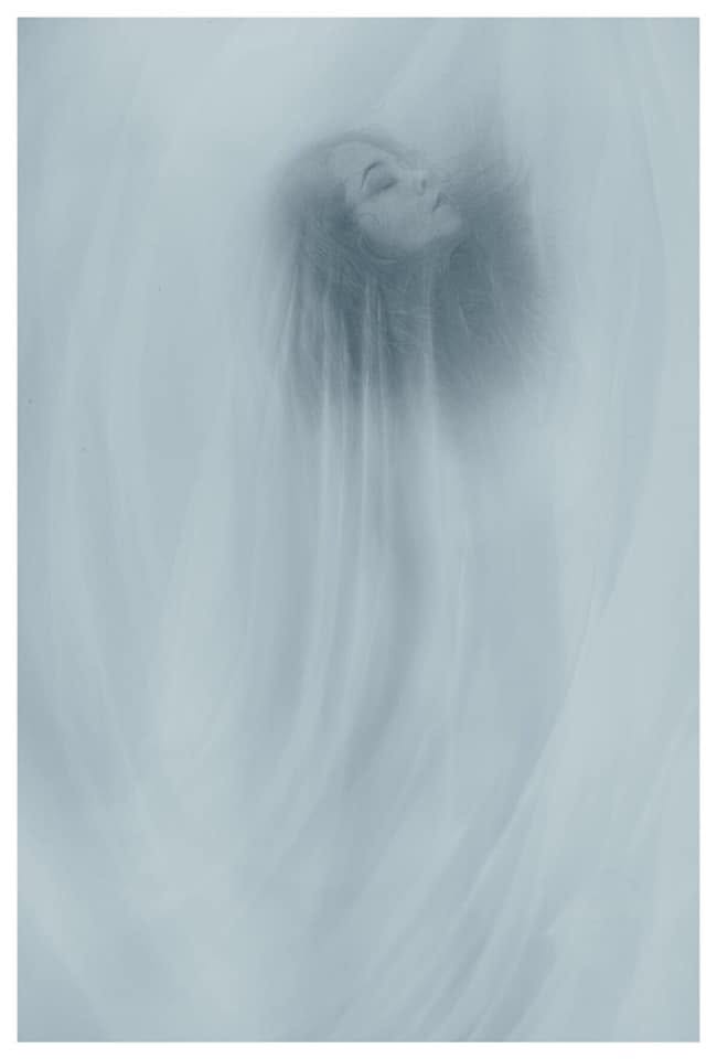 A person with long hair appears behind a semi-transparent curtain or fabric, with their face partially visible and eyes closed.