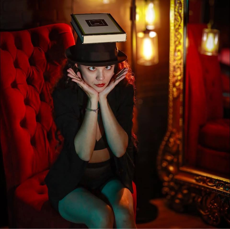 Woman with a book on her head, sitting on a red velvet chair in a dimly lit room.