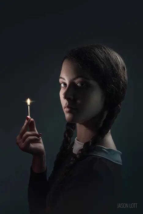 Young woman with braided hair holding a lit matchstick, dark background, serious expression.