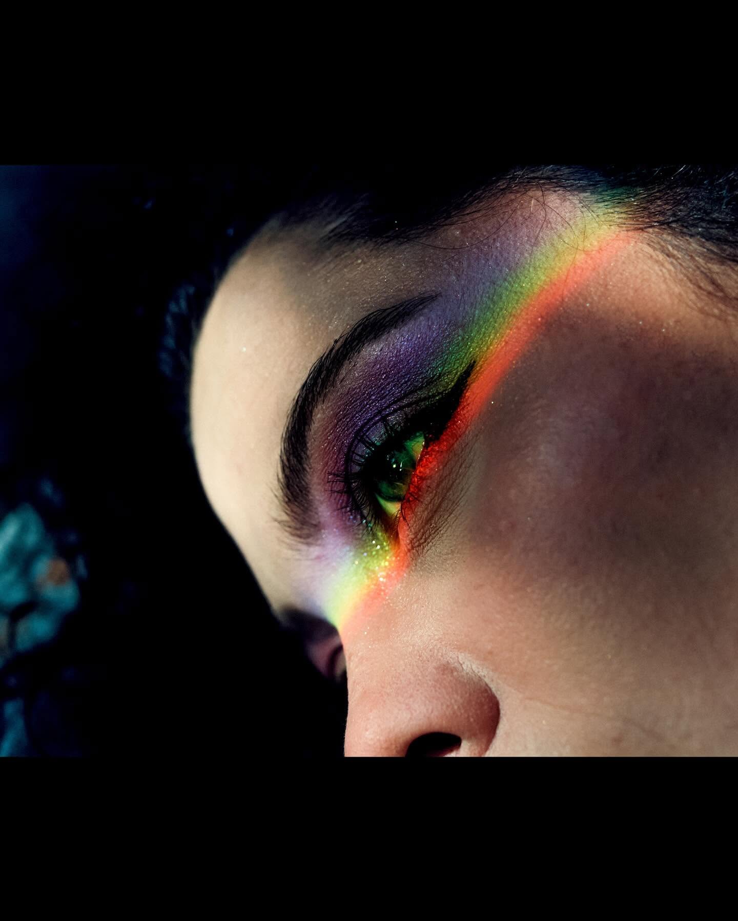 Close-up of a person's face with colorful makeup and a rainbow reflection on the eye, viewed sideways.