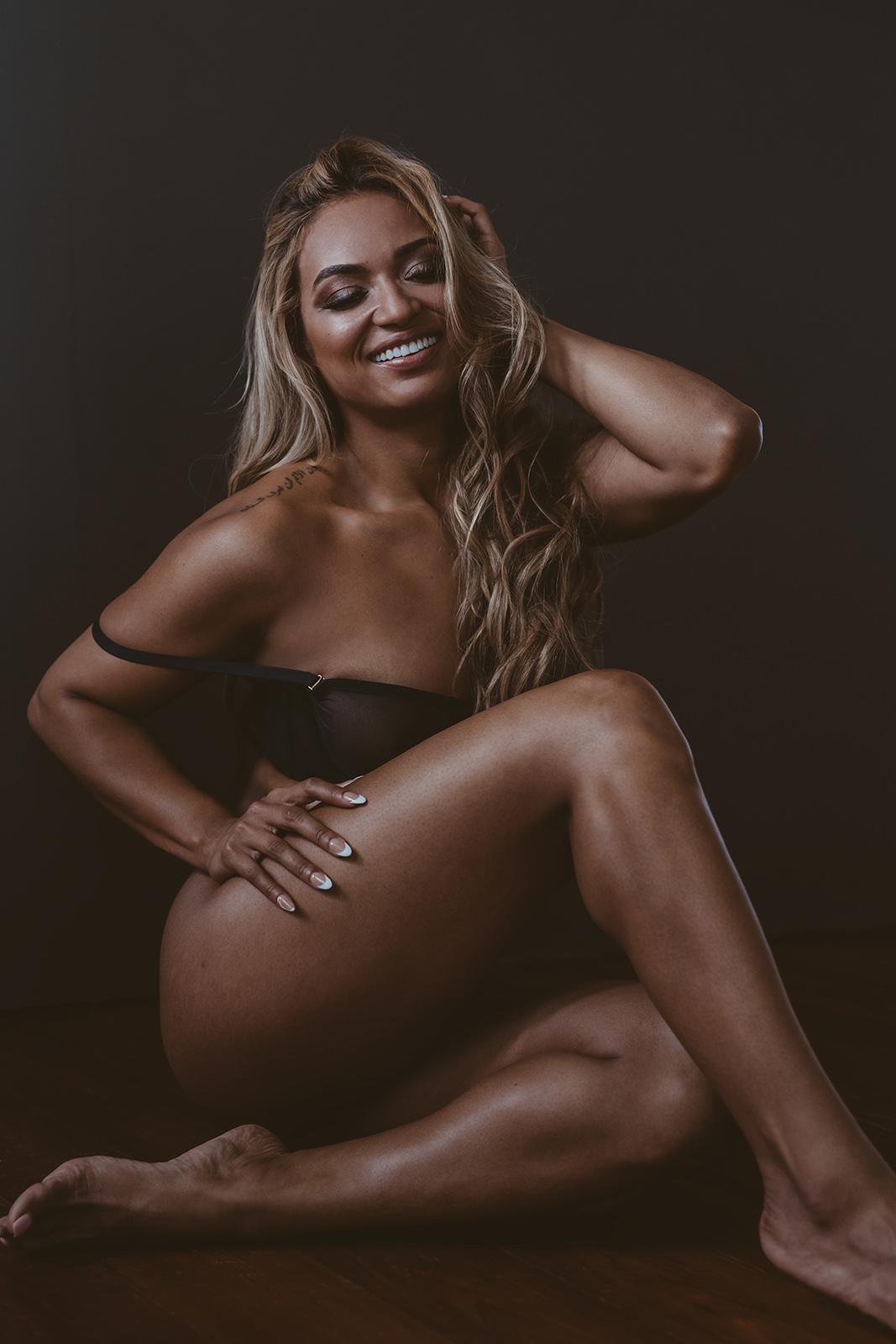 Smiling woman with long hair, sitting on the floor with one knee up, hand on her thigh, dark background.