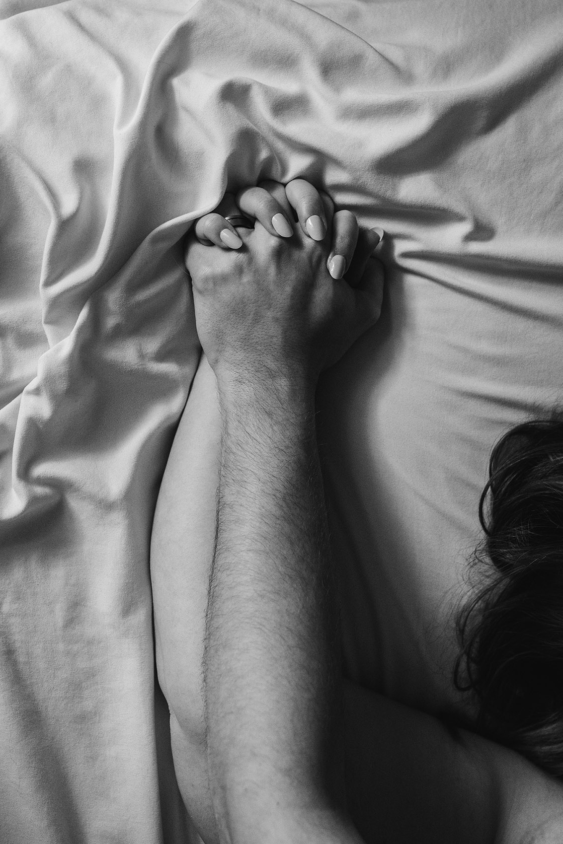 Person's hand and arm resting on a bed with crumpled sheets, black and white photo, partial view.