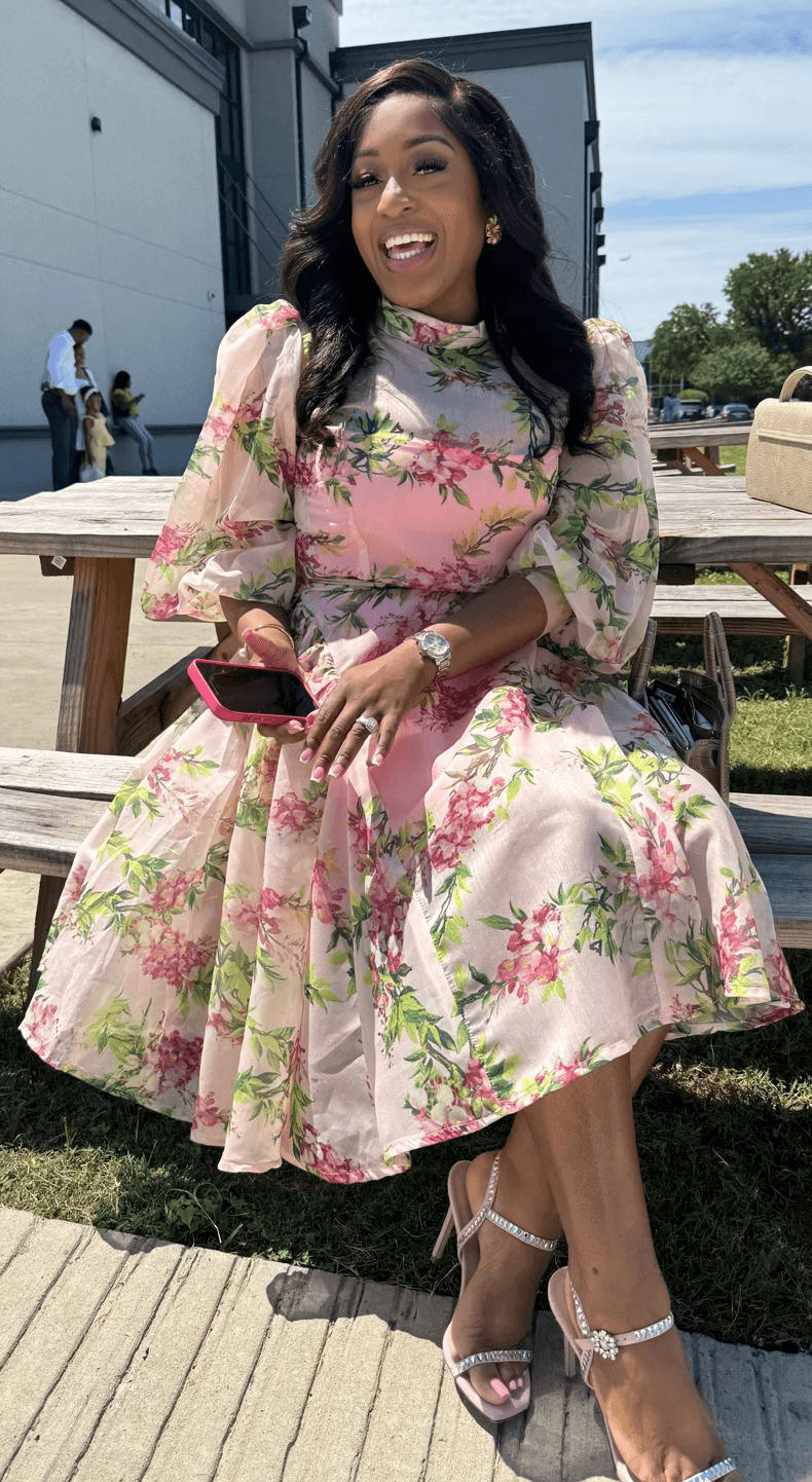 Woman in floral dress sitting outdoors, smiling, holding a phone, with people and trees in background.