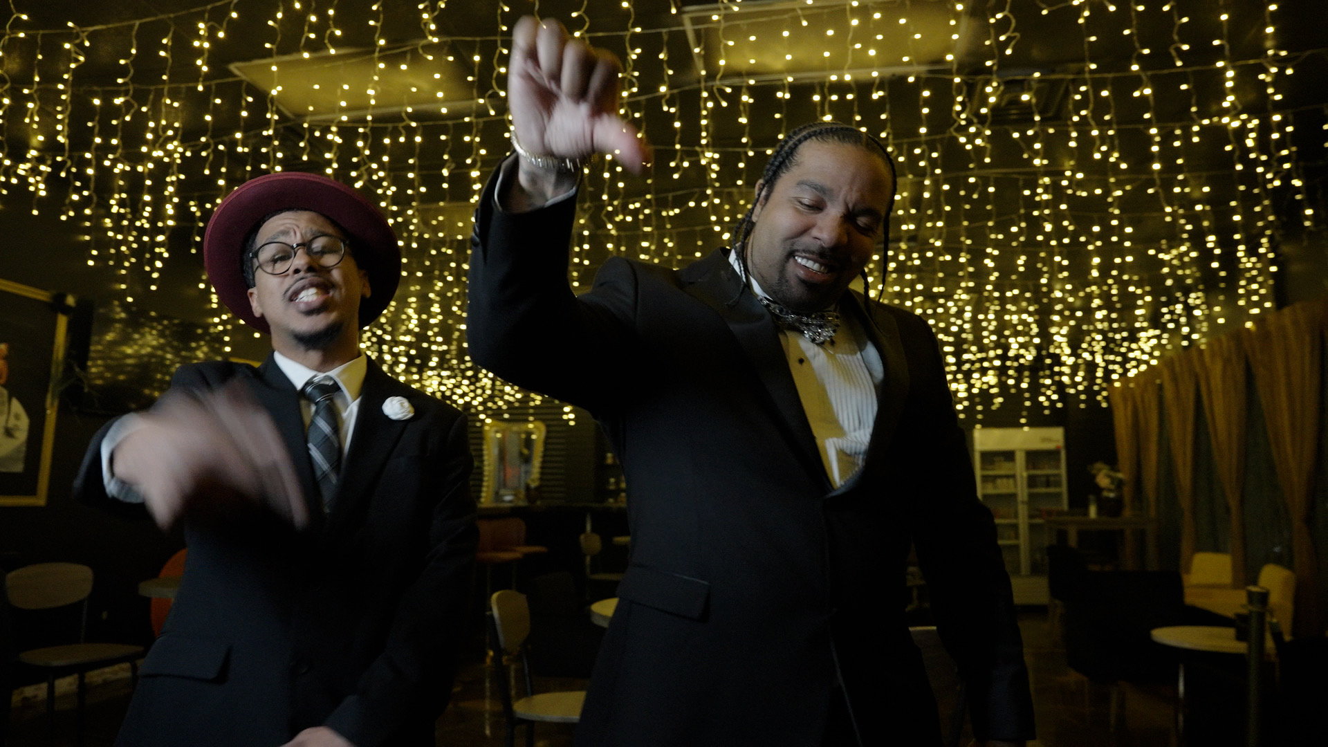 Two men in suits and hats dancing in a decorated room with hanging string lights, one raising his hand.