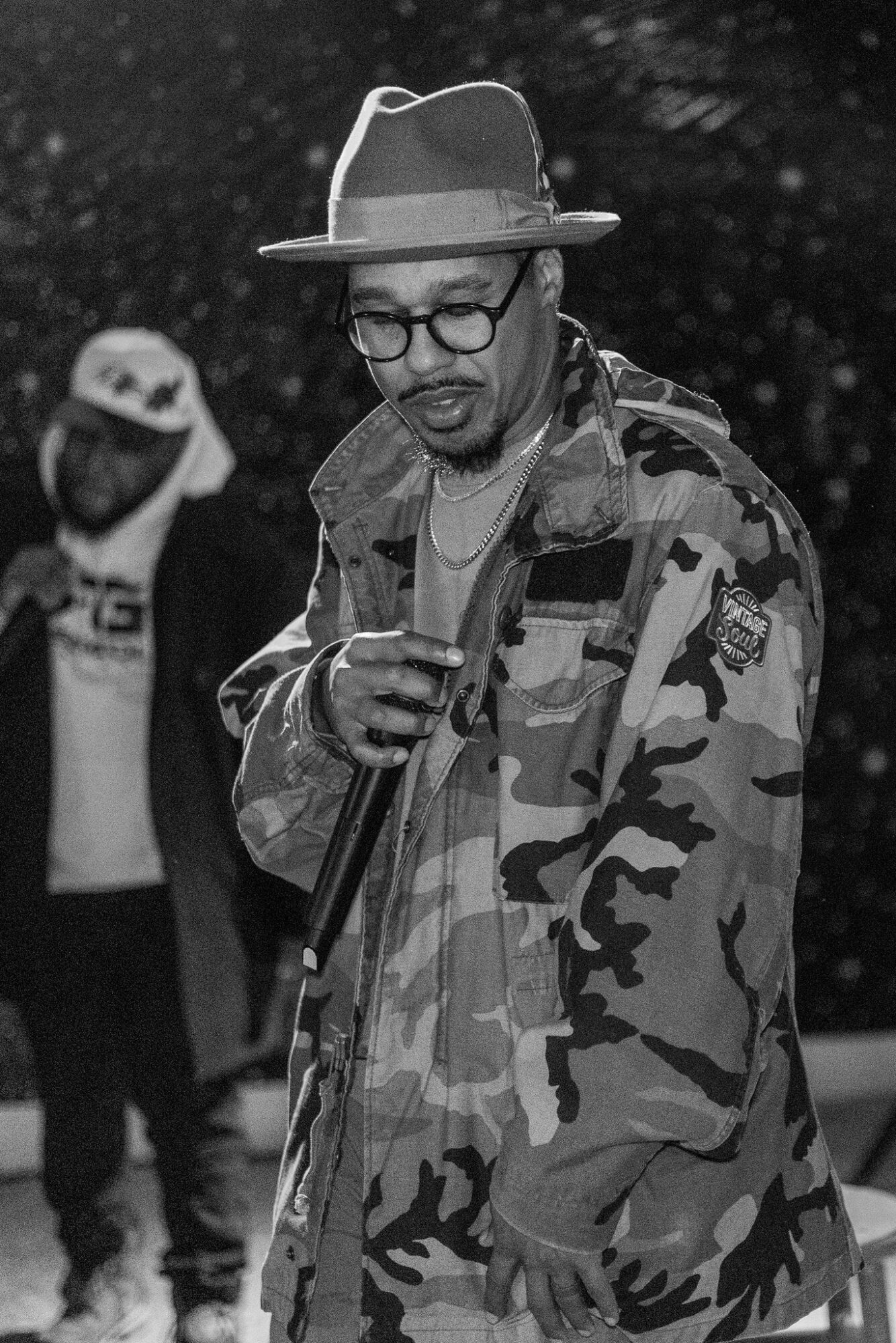 Man wearing glasses, hat, camouflage jacket, holding a microphone, with another person in background outdoors.