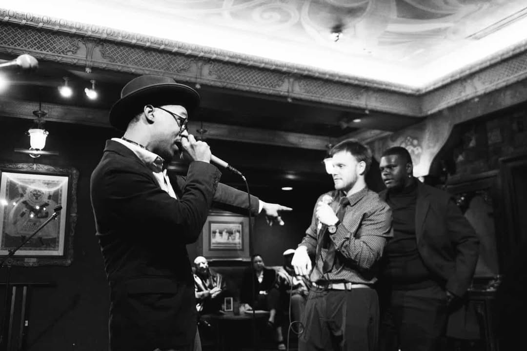 Three men in a room with ornate ceiling, one singing into a microphone, others watching, framed pictures on walls, dim lighting.
