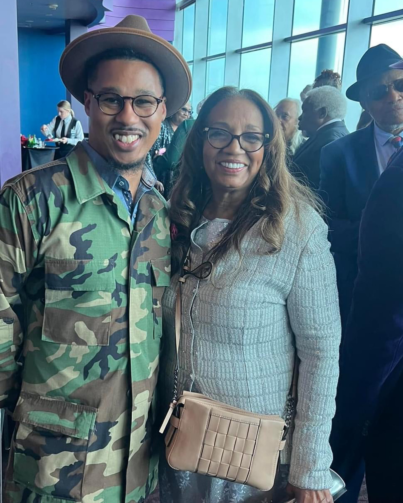 Two people smiling at an indoor event, one wearing a camouflage jacket and hat, the other in a light-colored blazer, with large windows behind them.