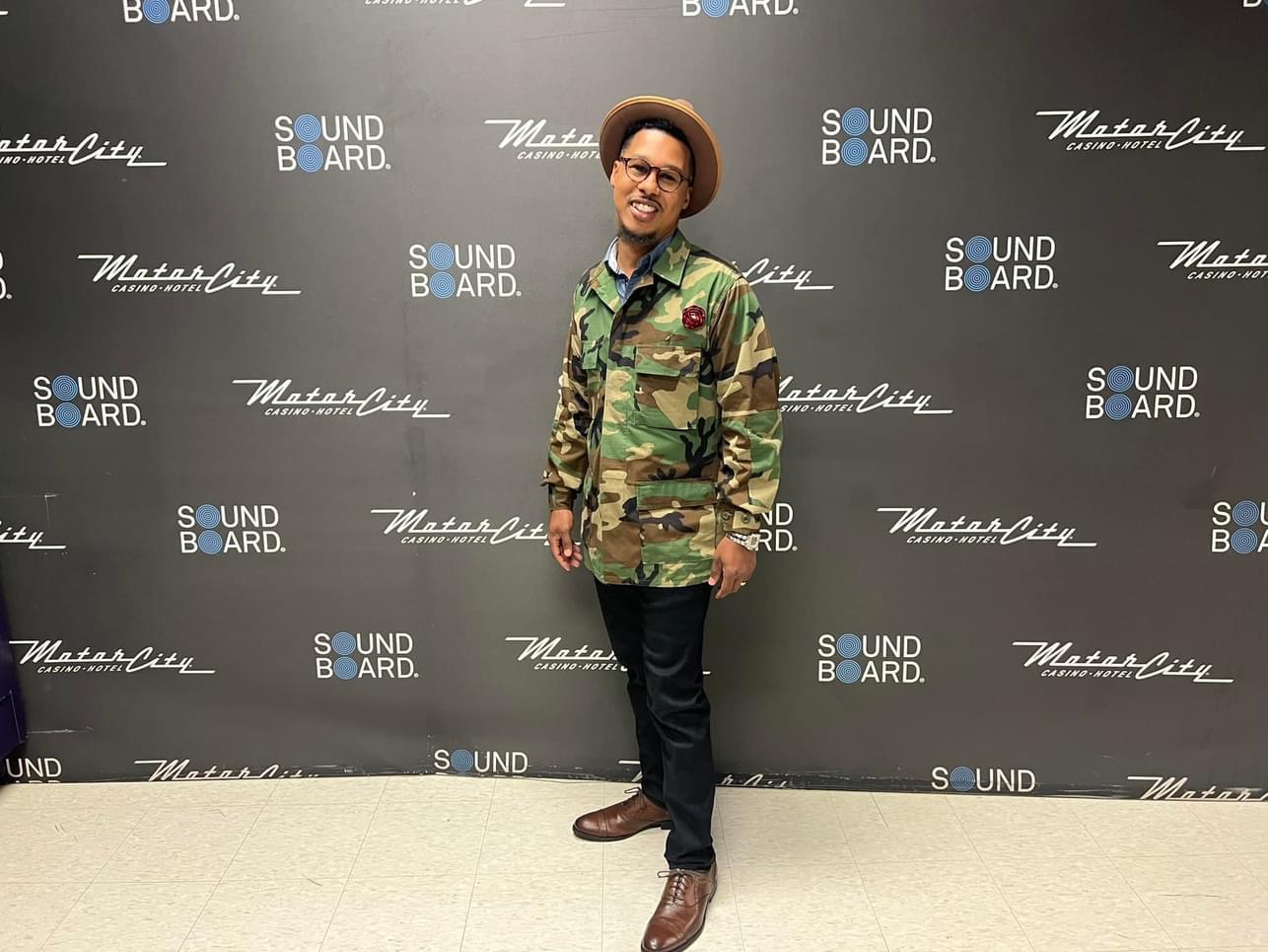 Person standing in front of a black backdrop with logos, wearing a camouflage jacket, black pants, and brown shoes.