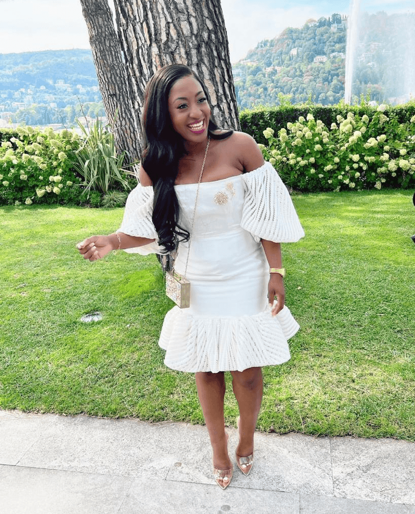 Smiling woman in white dress standing outdoors near a tree and bushes, with a fountain and landscape in background.
