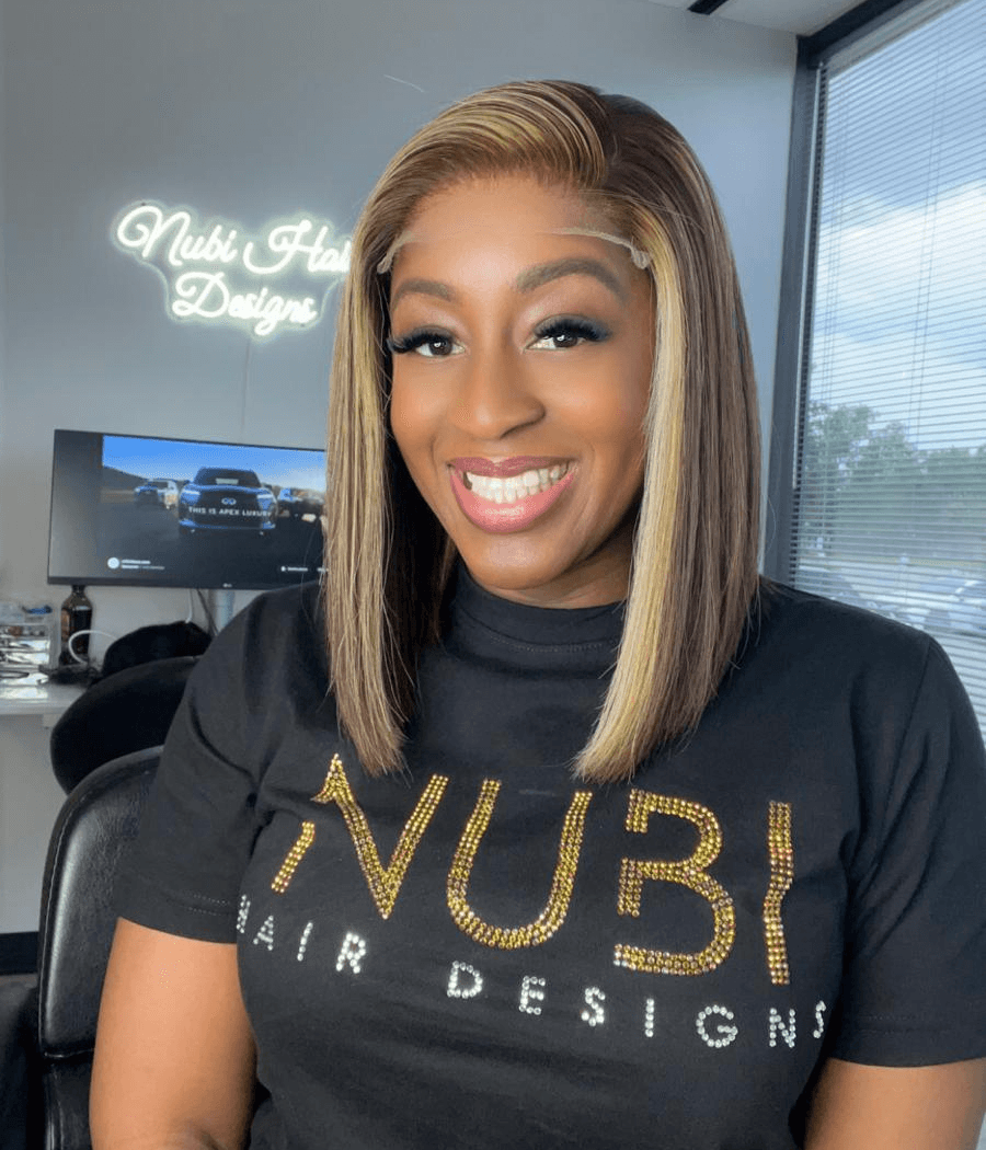 Woman with shoulder-length blonde hair smiling, wearing a black T-shirt with gold lettering, in an office setting.