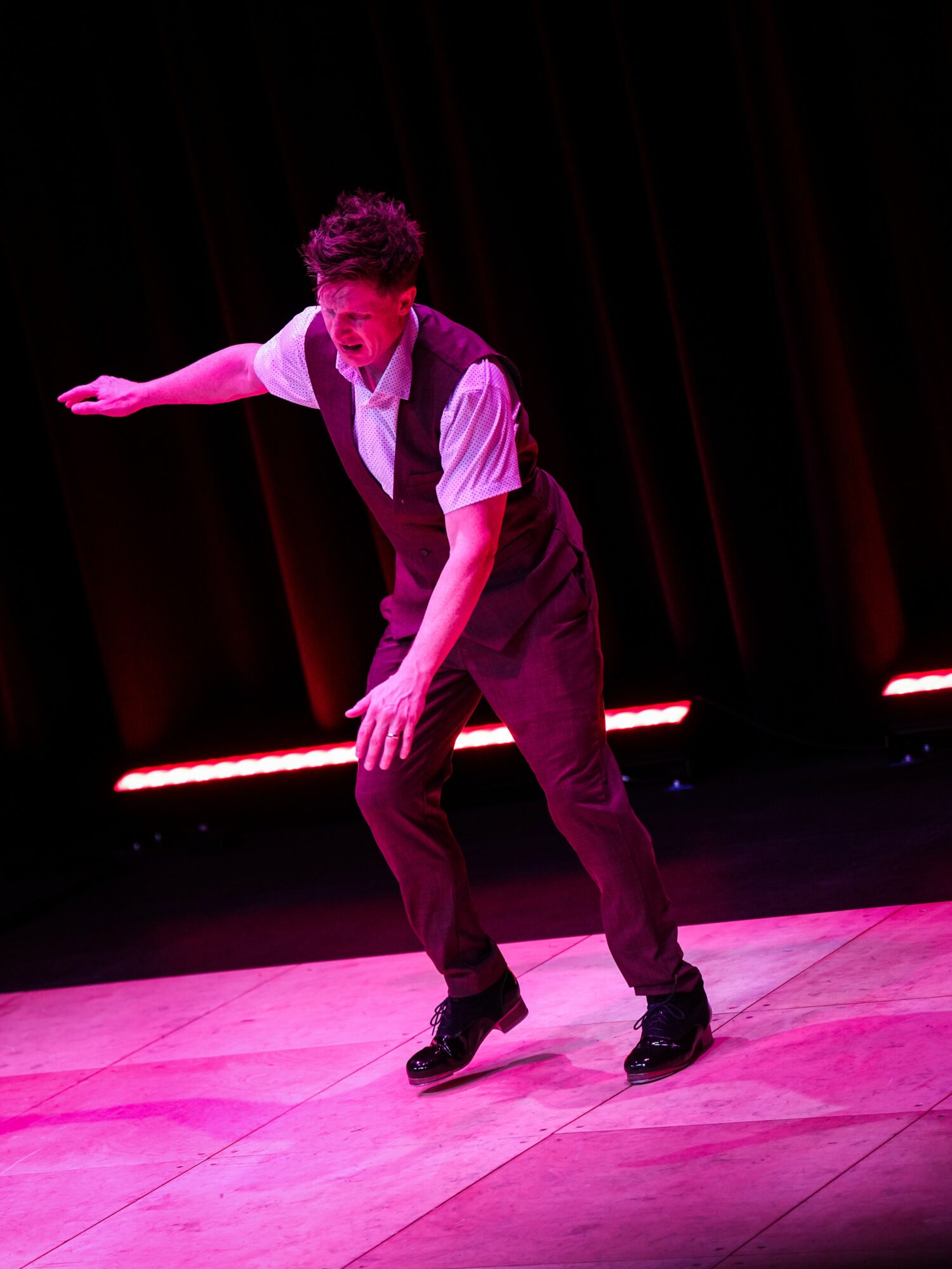 Person dancing on stage with dark background, wearing a white shirt and dark pants, in a dynamic pose.