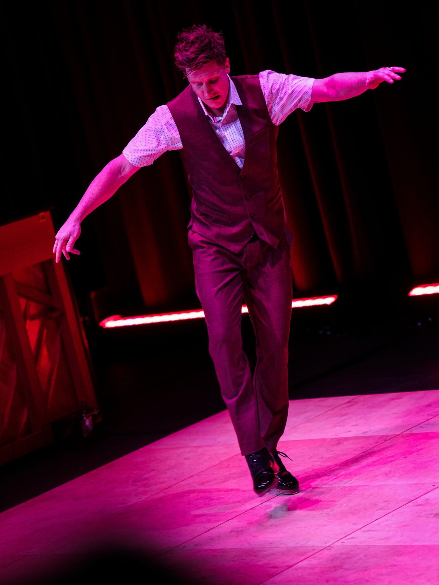 Performer in dark vest and pants, arms extended, on stage with red lighting and curtains behind.