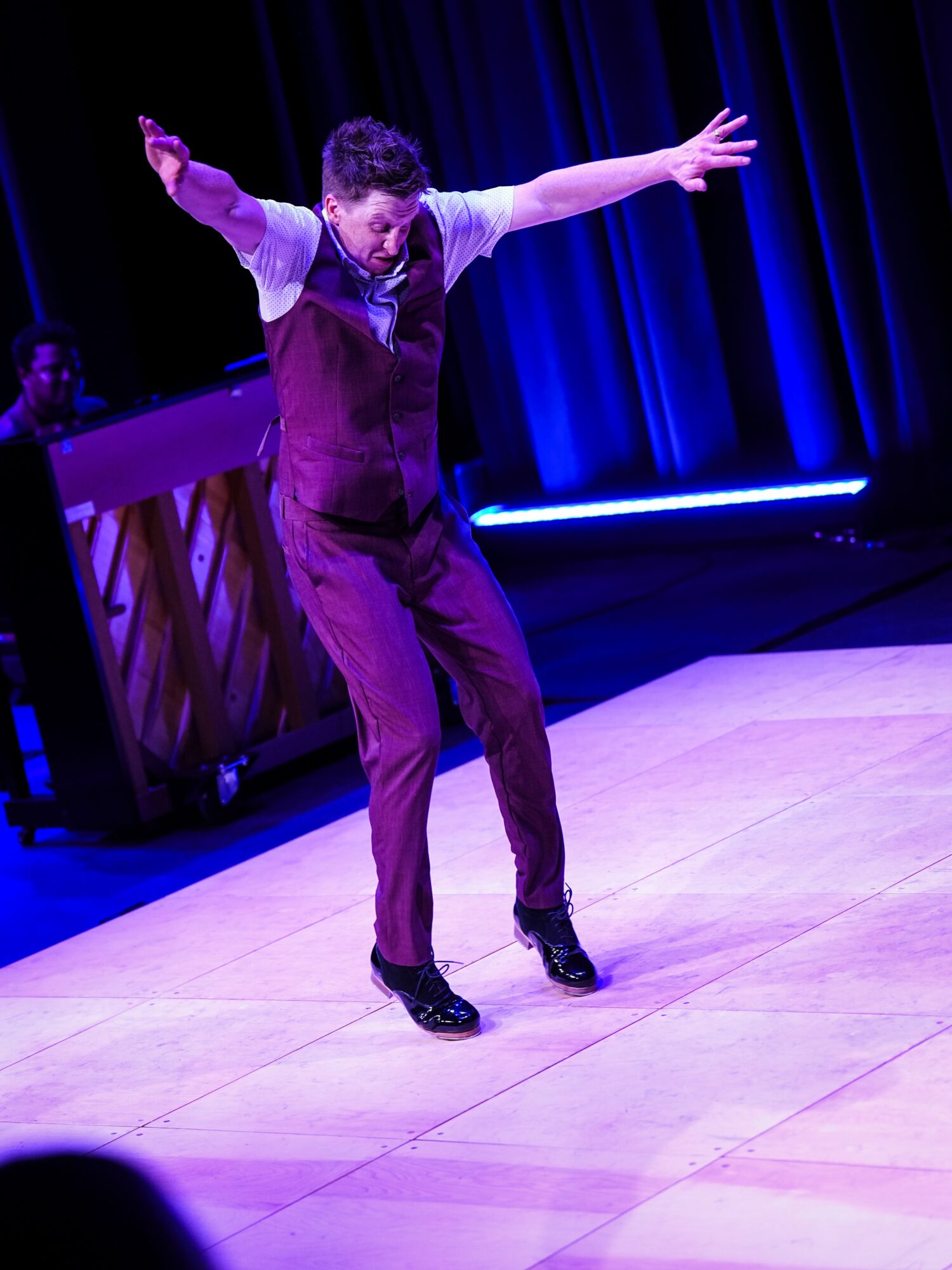 Person in a purple vest and pants dancing on stage with arms outstretched, dark background, stage lighting, wooden floor.