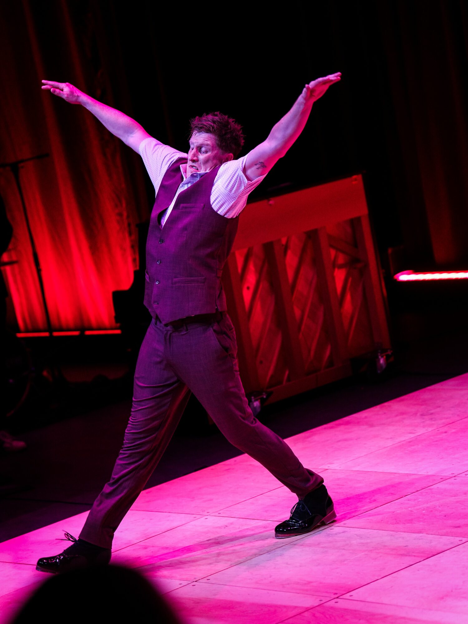 Person dancing on stage with arms raised, wearing a vest and pants, with a red curtain and piano in background.