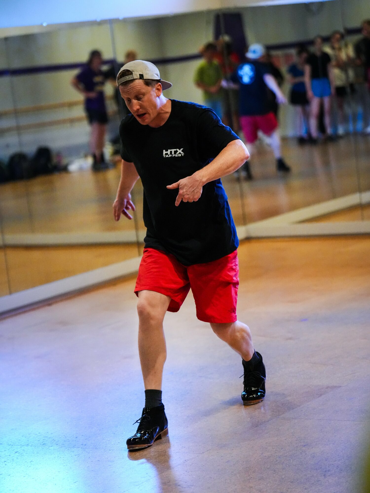 Young person in black shirt, red shorts, and black shoes skating indoors, with a mirror and people in the background.