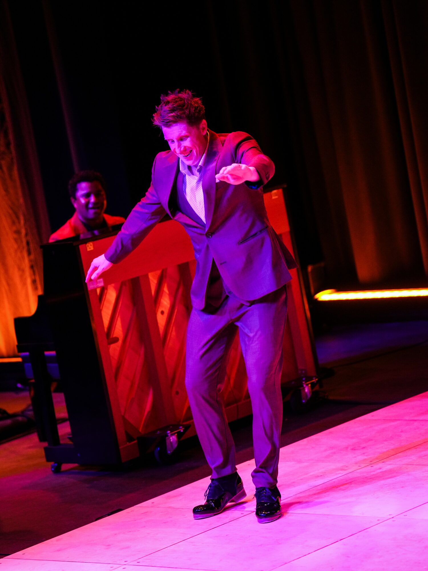Person in a suit dancing on stage with a piano and another person in the background, colorful stage lighting.