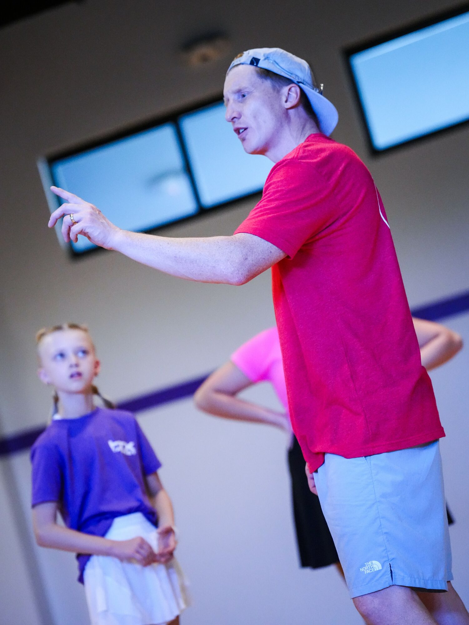 Young man in red shirt and cap gesturing while talking to children in gymnasium.
