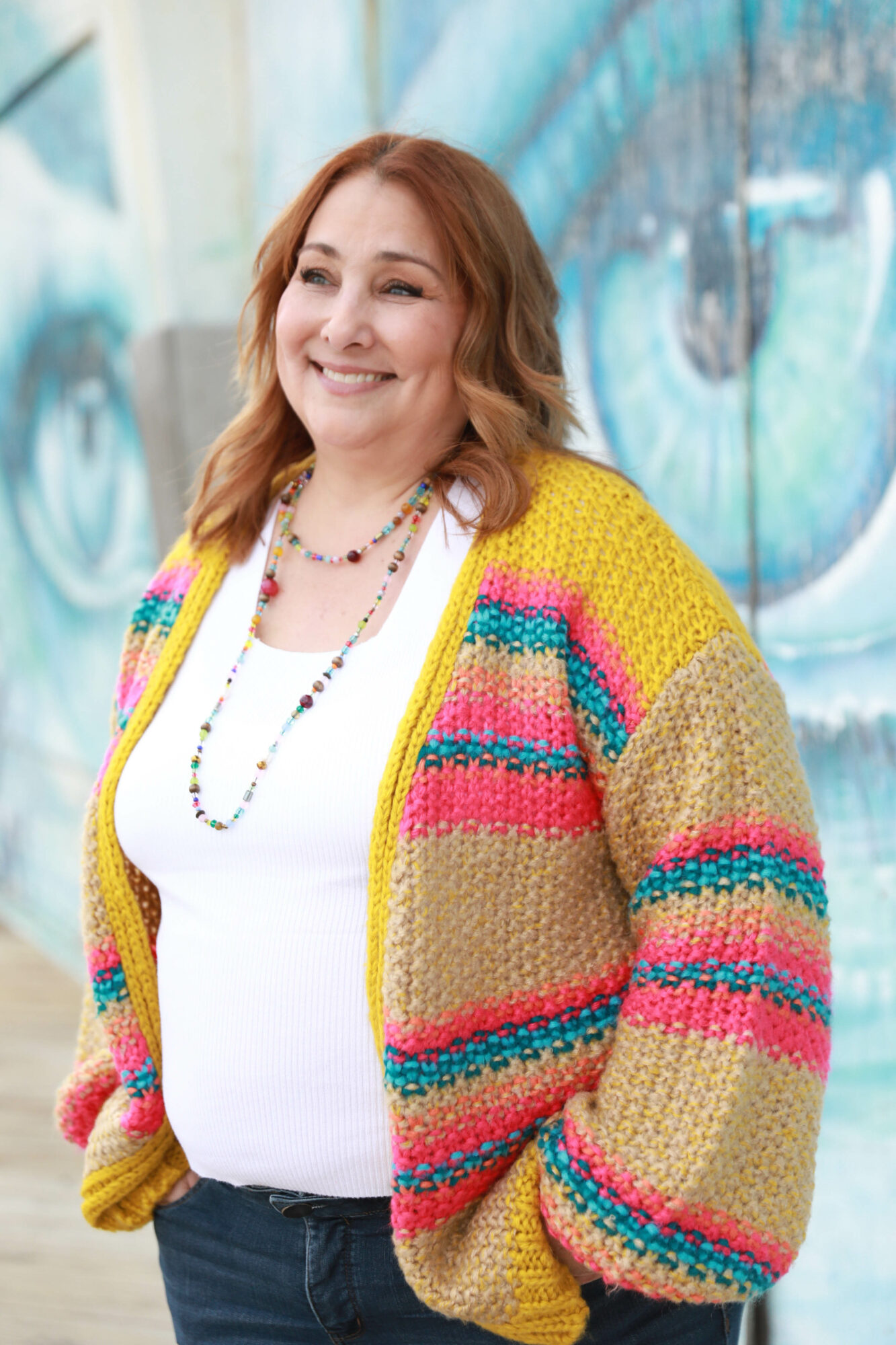 Smiling woman with wavy red hair wearing a colorful striped cardigan and white top, standing outdoors with a graffiti wall background.