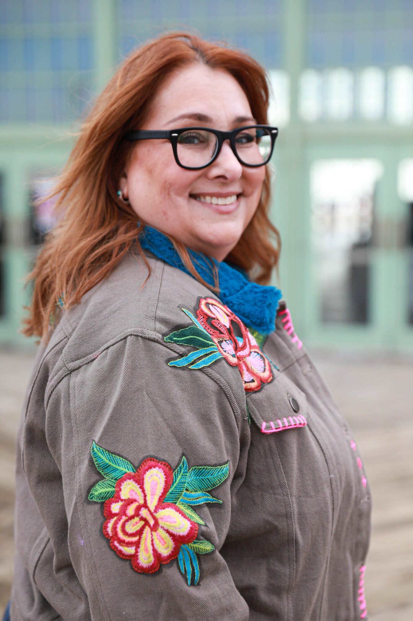 Woman with glasses and red hair smiling outdoors, wearing a gray jacket with floral embroidery.