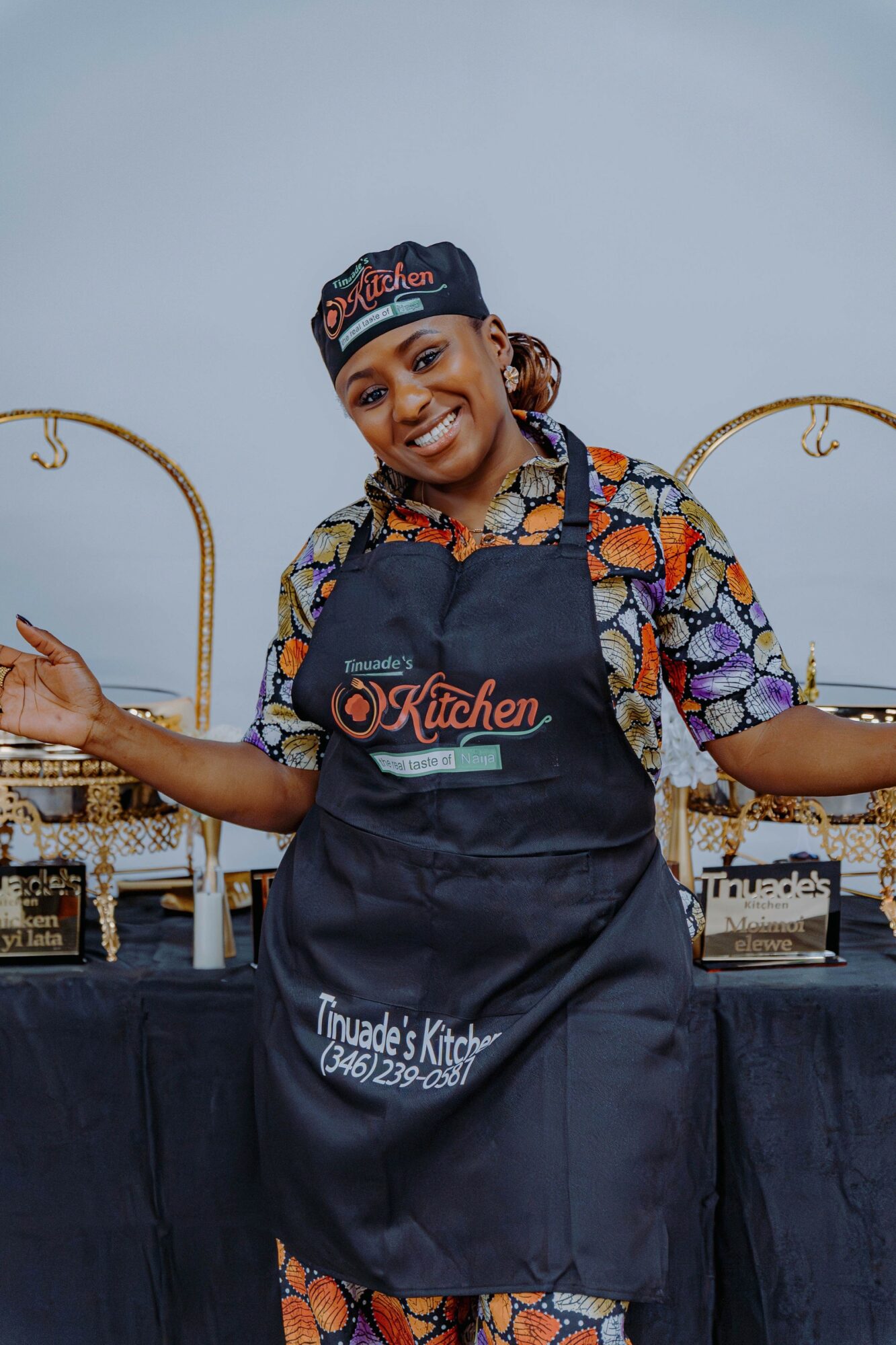 Smiling woman in colorful shirt and apron stands behind a table with baked goods, arms outstretched, in a bakery setting.