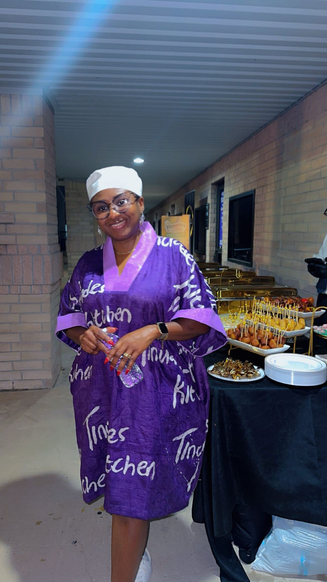 Woman in purple dress and white headscarf standing next to a table with food, smiling, in an indoor setting.