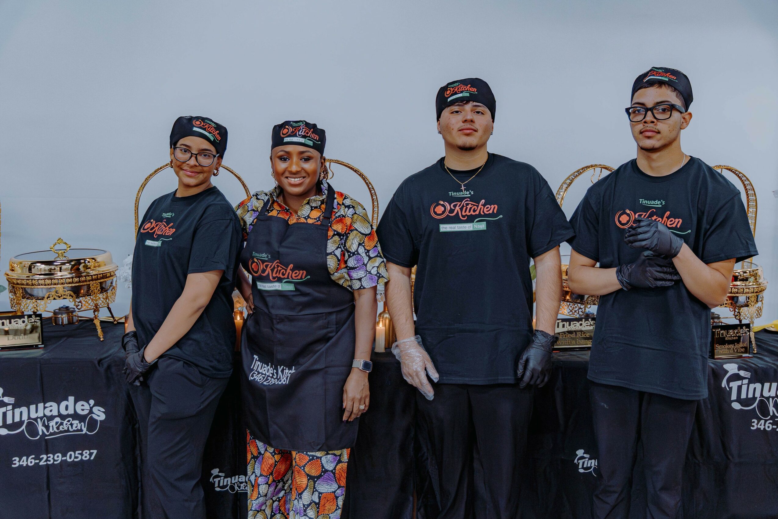 Four people stand in front of a table with food, wearing matching uniforms and aprons, smiling at the camera.