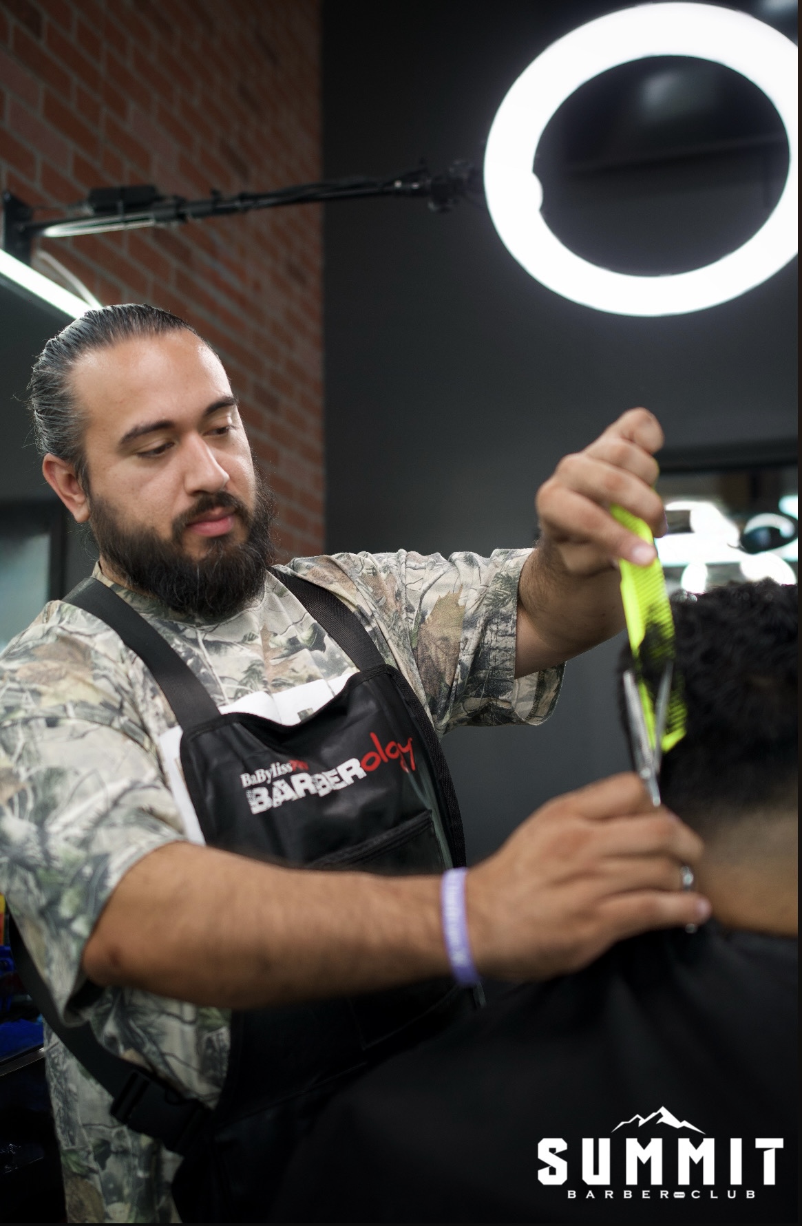 Barber with beard and apron cutting a customer's hair in a modern salon, illuminated by a ring light, brick wall background.