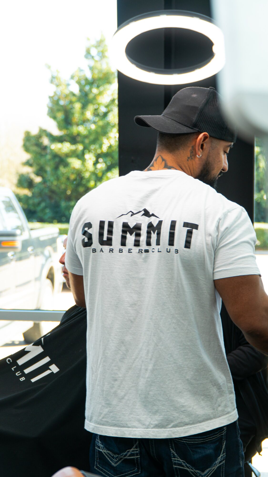 Person wearing a white T-shirt with 'SUMMIT' text, black cap, and beard, standing indoors near a circular light fixture.
