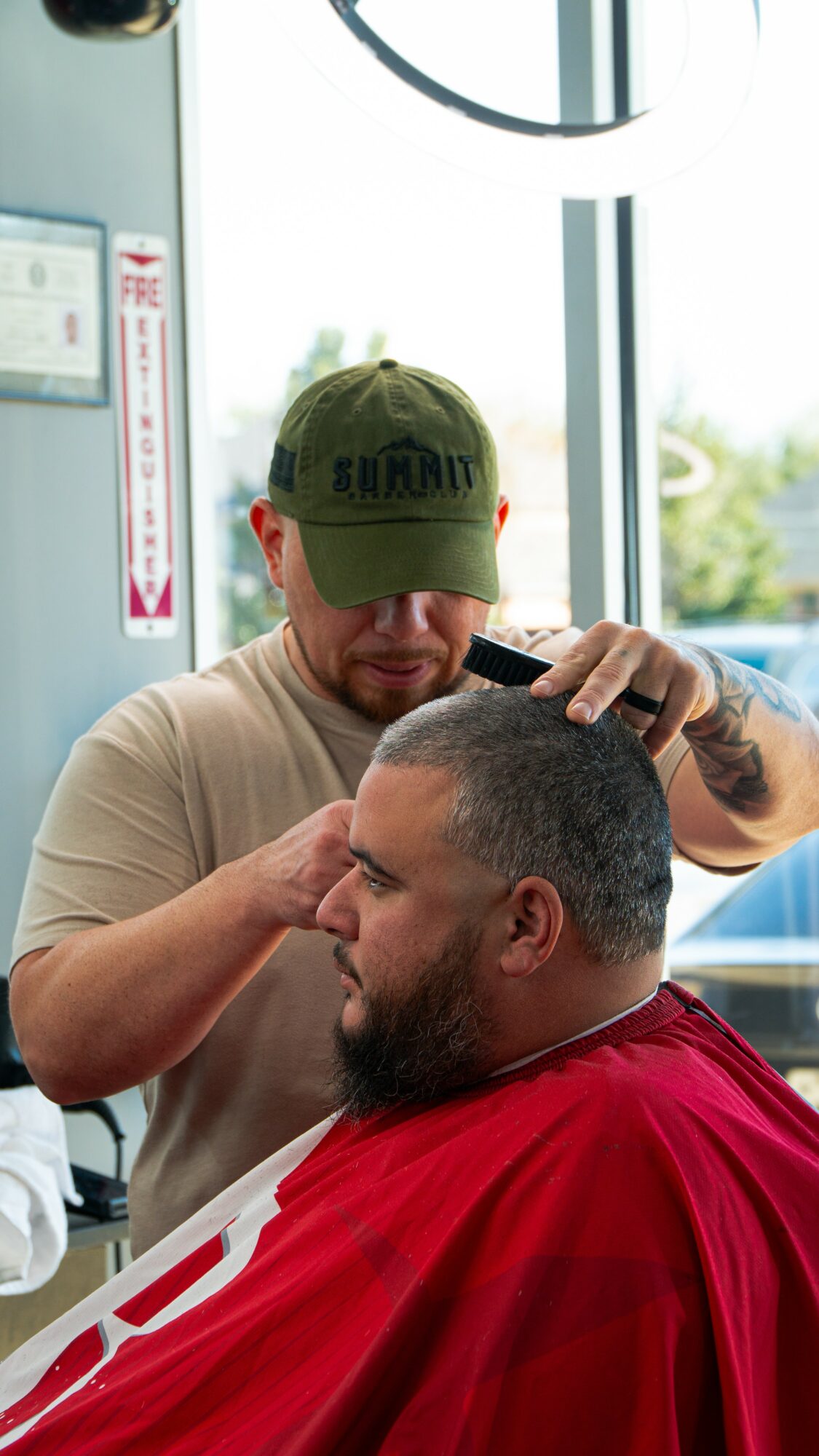 Barber trims a man's hair in a barbershop, with a window and natural light in the background.