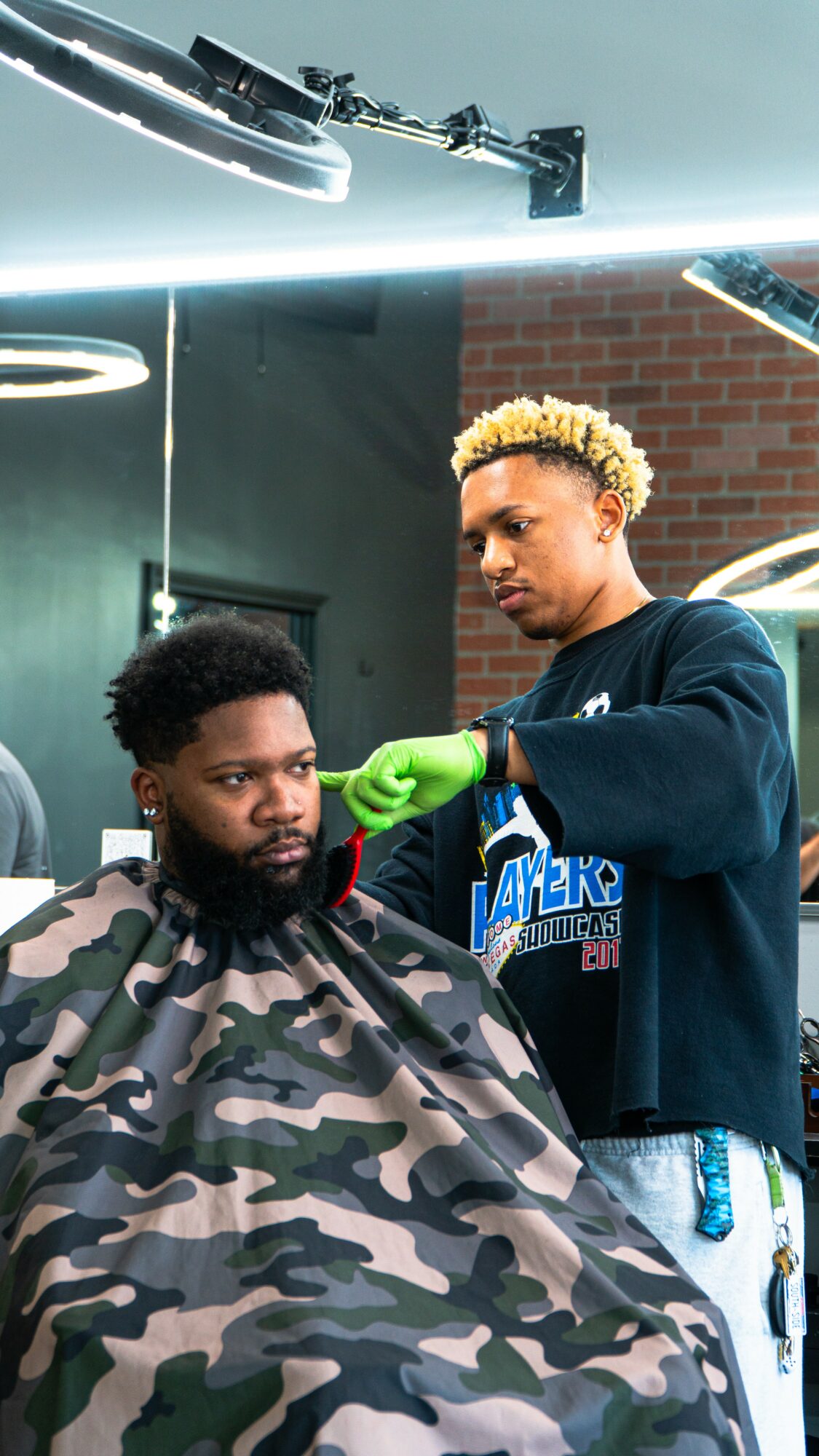 Barber cutting a man's hair in a salon with brick wall background, using green gloves, with salon equipment overhead.