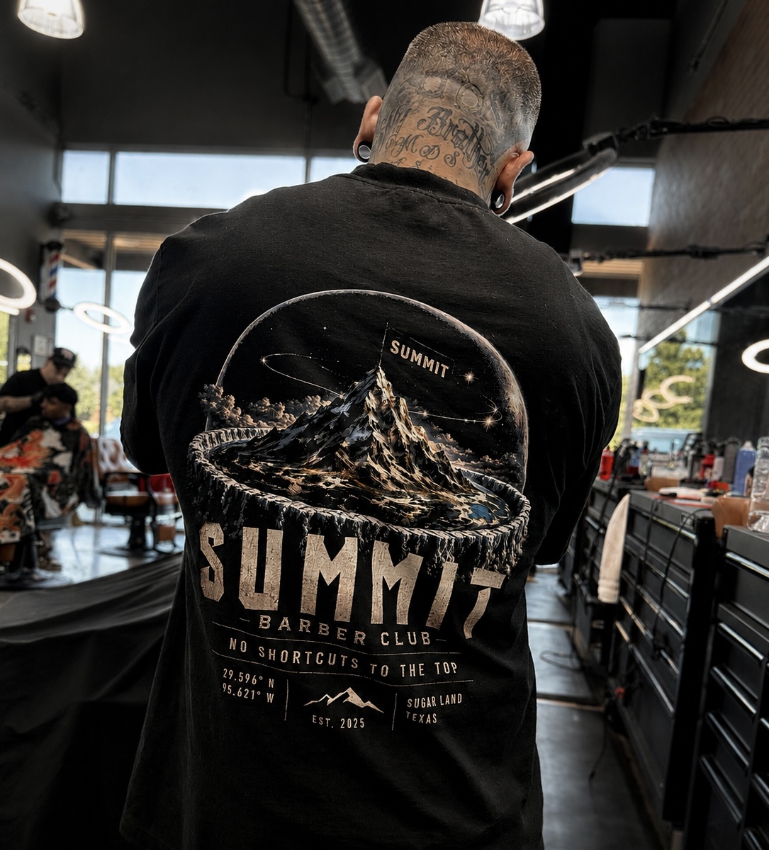 Person with a shaved head wearing a black jacket with mountain and summit design, inside a modern cafe or restaurant.