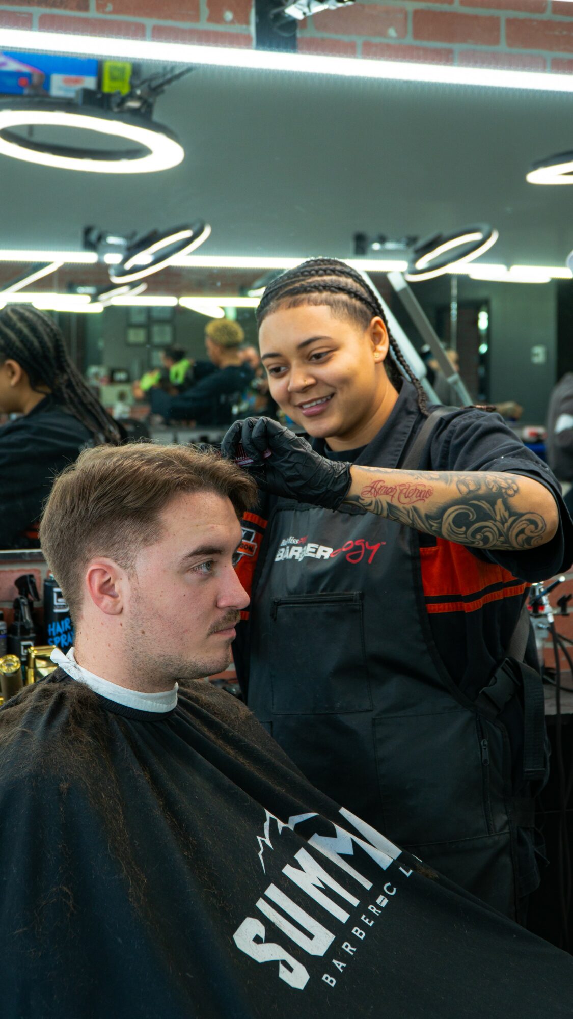 Barber styling a man's hair in a barbershop with bright lighting and mirrors, people in the background, smiling woman with tattoos.