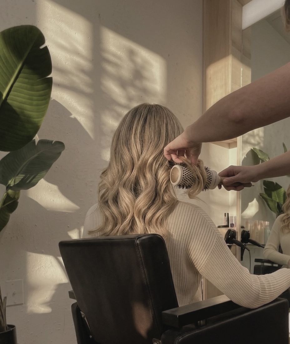 Person with long, wavy hair sitting in a chair, getting styled by a hairdresser in a well-lit room with plants.