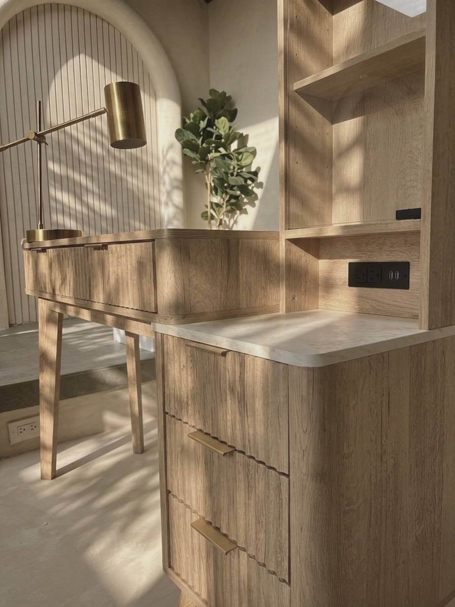 Wooden desk with drawers and shelves, sunlight casting shadows, a potted plant, and a brass lamp in the background.