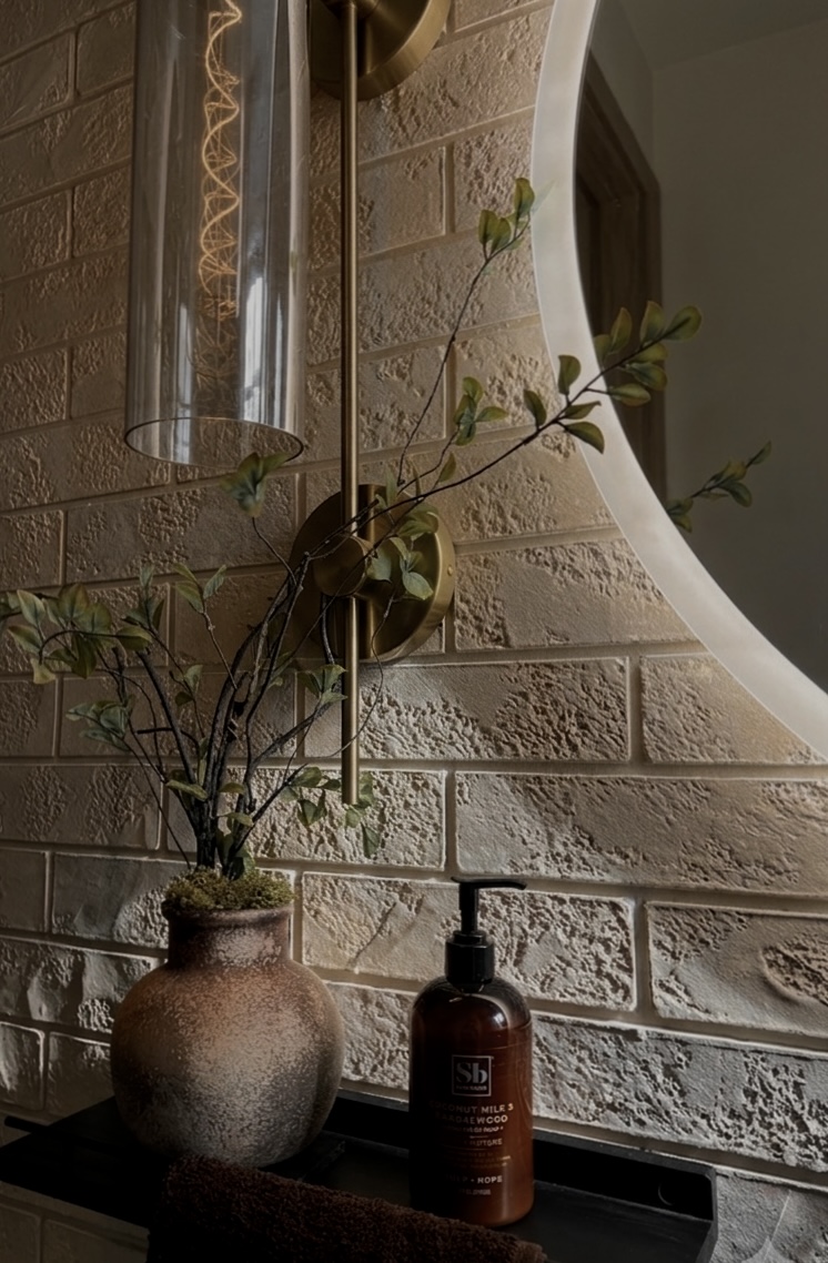 Vase with plant, soap dispenser, and round mirror on a brick wall, illuminated by warm lighting.
