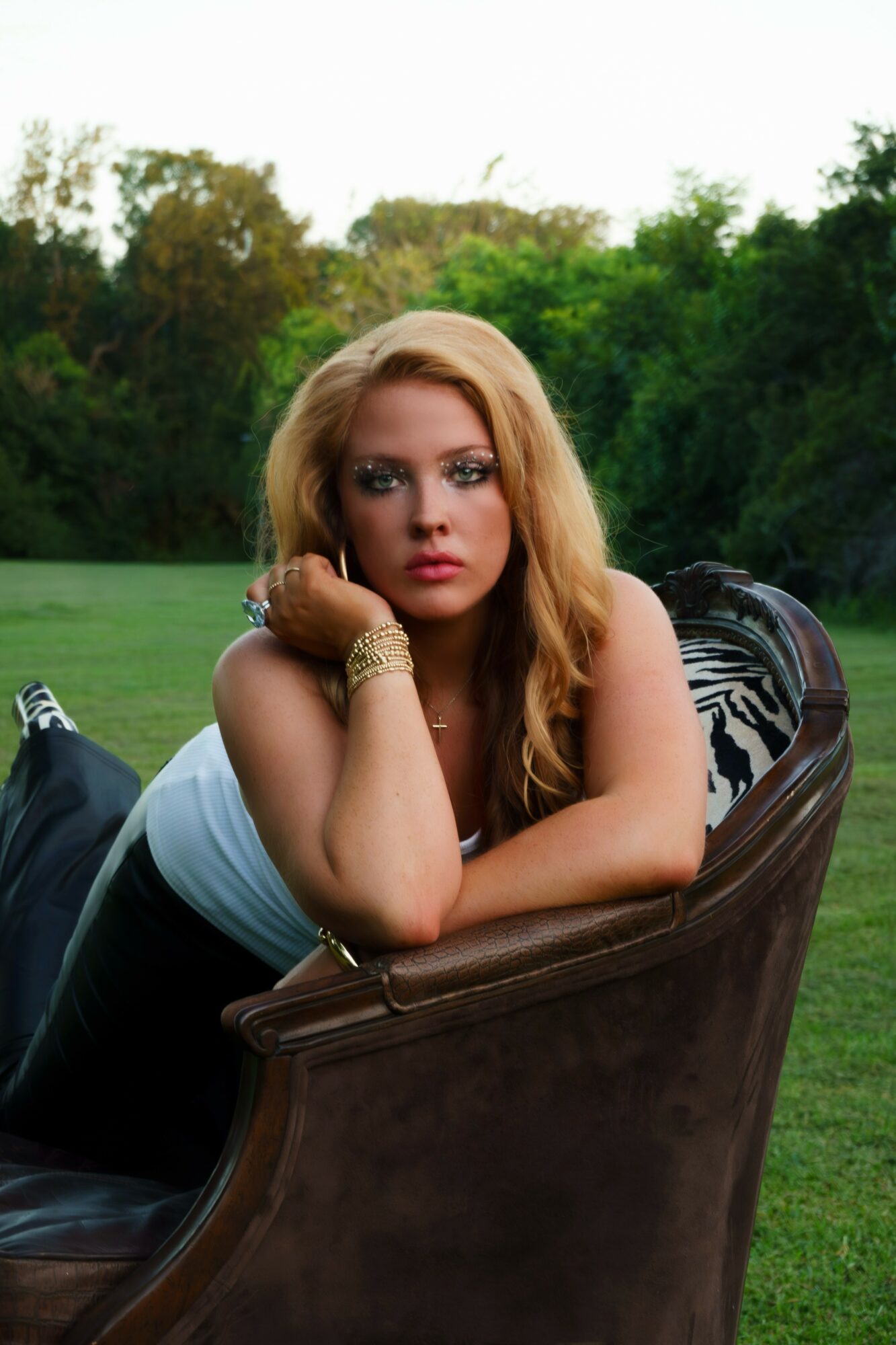 Woman with long blonde hair resting her head on her hand outdoors, sitting on a chair, with trees in the background.