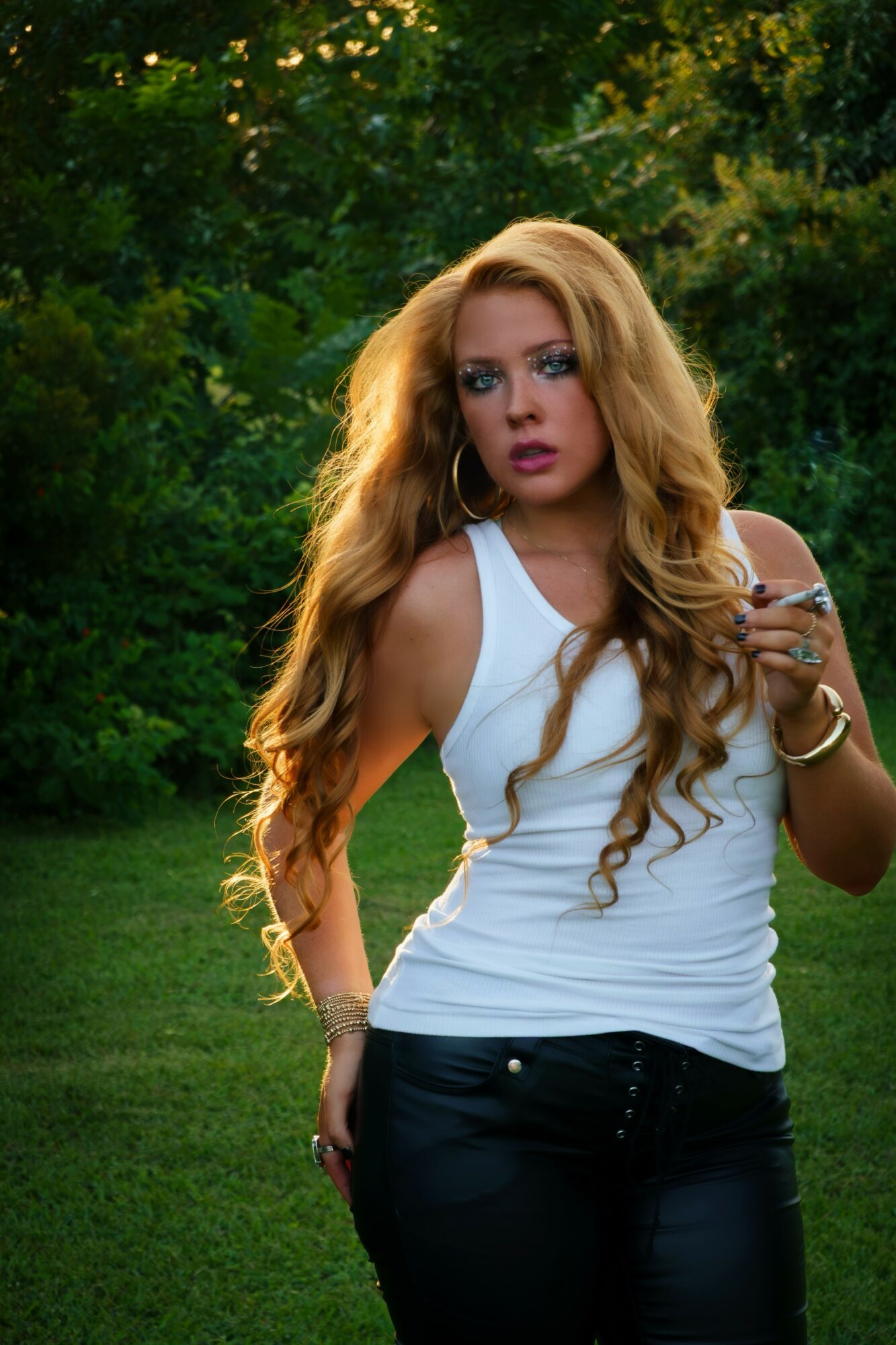 Young woman with long, wavy hair wearing a white sleeveless top and dark pants outdoors, holding a phone.