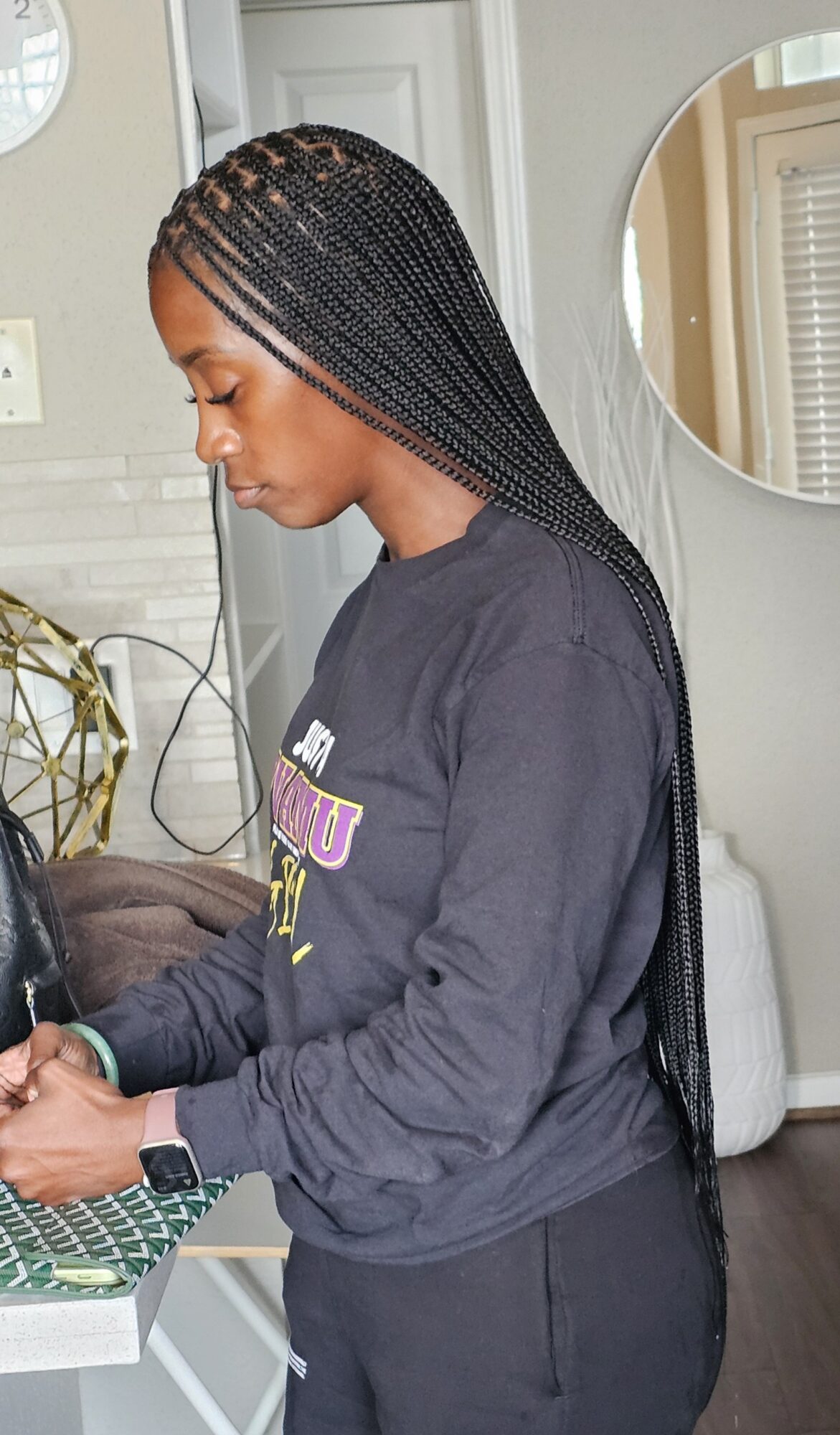 Young woman with braided hair looking down, wearing a dark sweatshirt, in a room with a mirror and door in background.