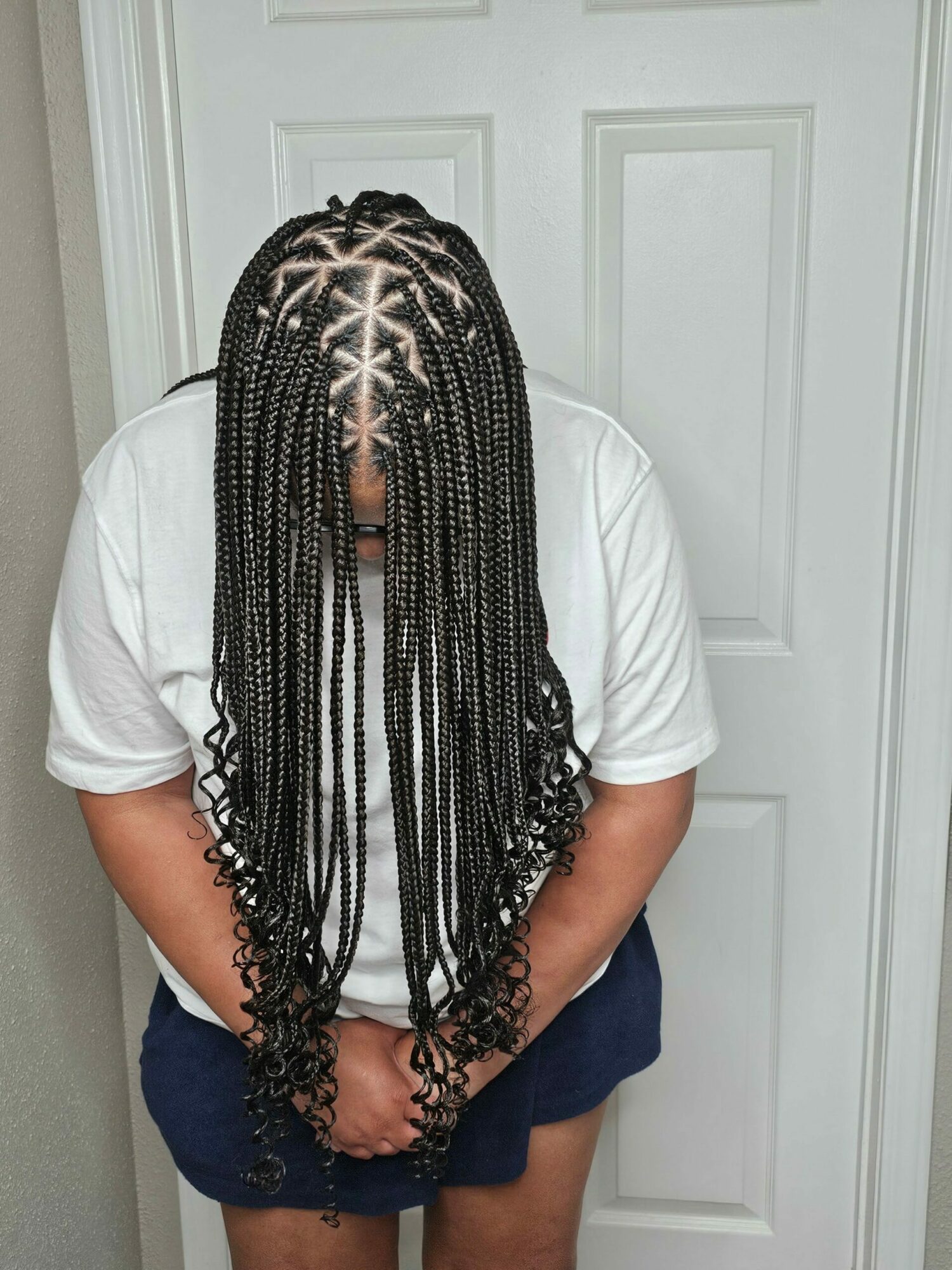 Person with long braided hair wearing a white shirt and dark shorts, standing against a white door.