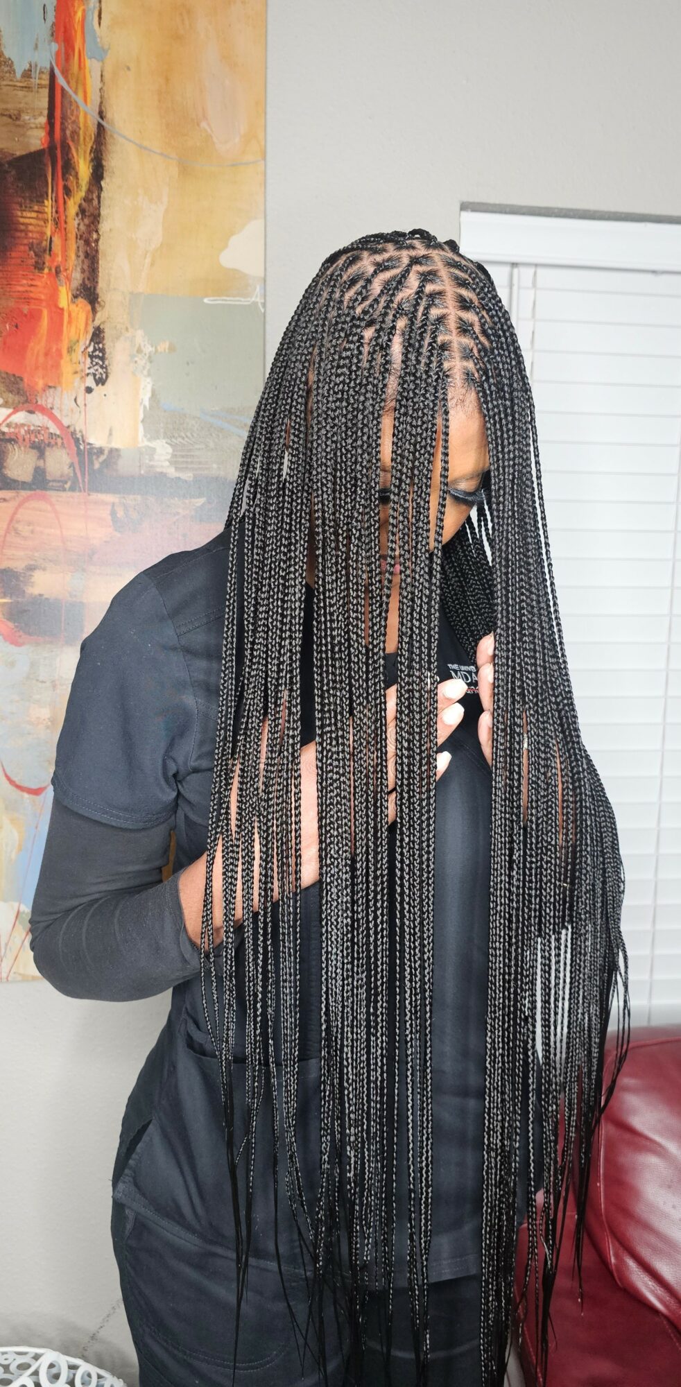 Person with long braided hair wearing a dark jacket, standing indoors near a window and a red chair.