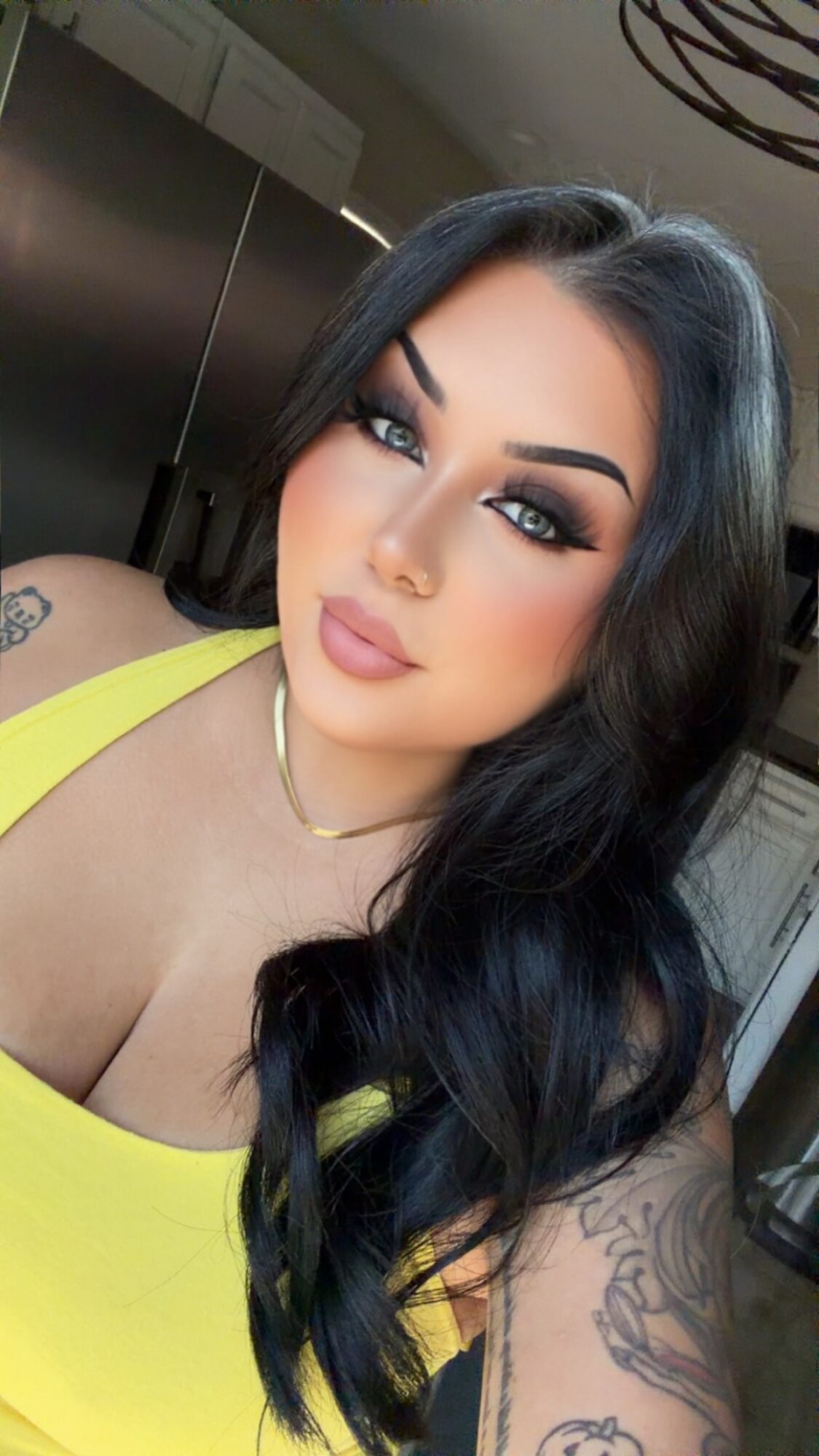 Woman with long dark hair, makeup, and a yellow top, looking at camera indoors.